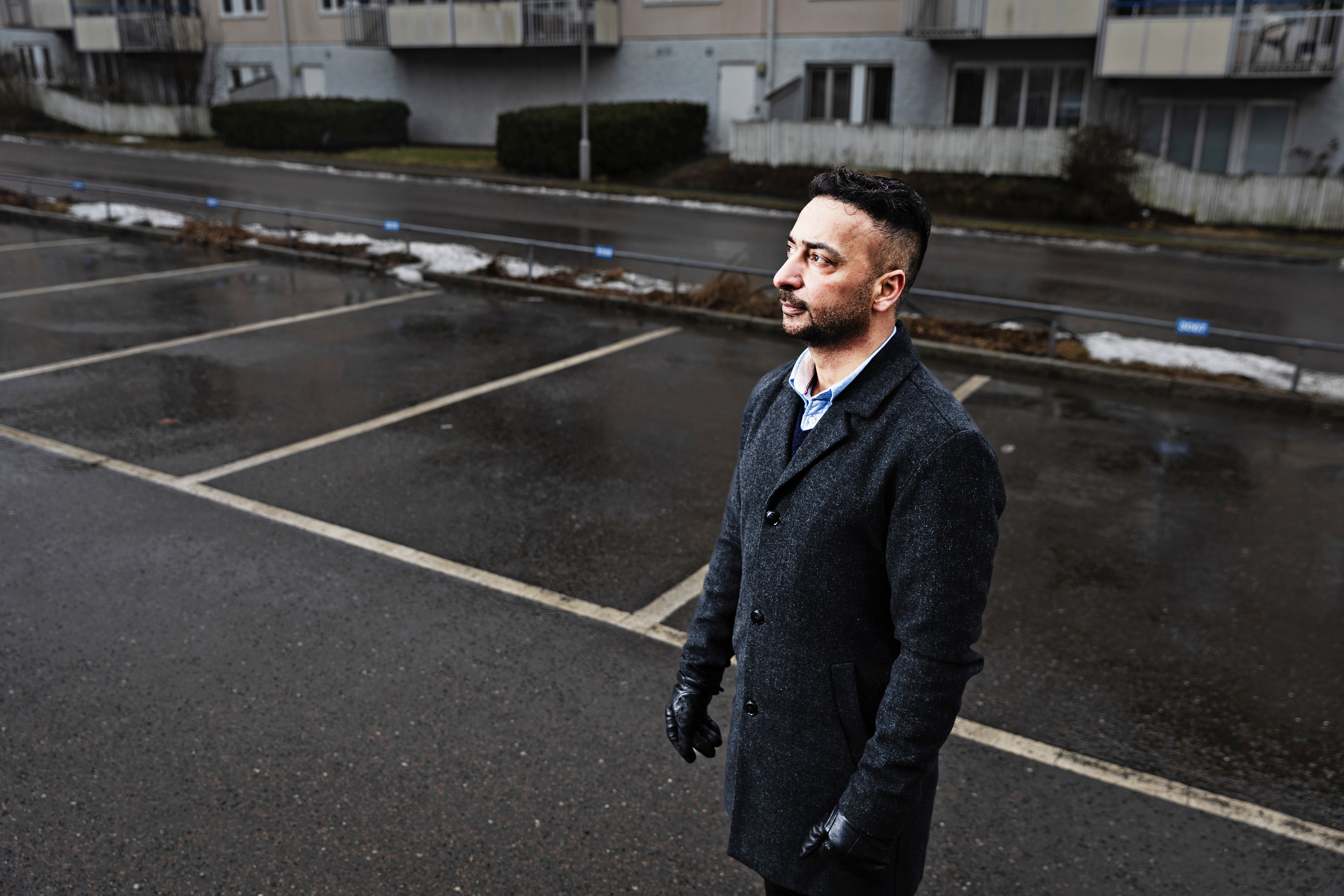 Rabie Al Rubeai med mörk rock och handskar står på en tom parkering. Bakom honom finns våt asfalt och en lägenhetsbyggnad.