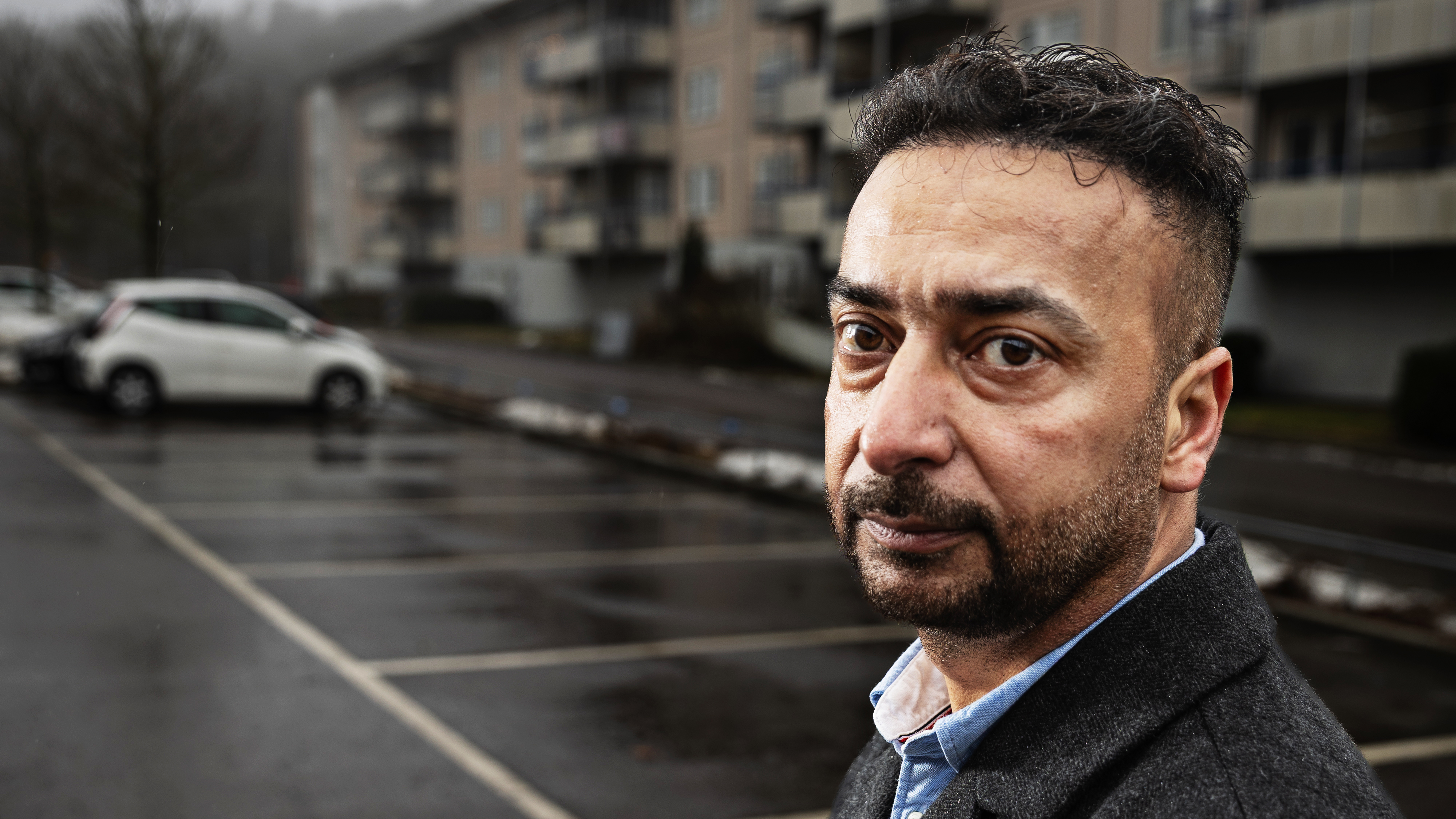 En man med kort mörkt hår och skägg står på en nästan tom parkering framför ett lägenhetshus en mulen dag.