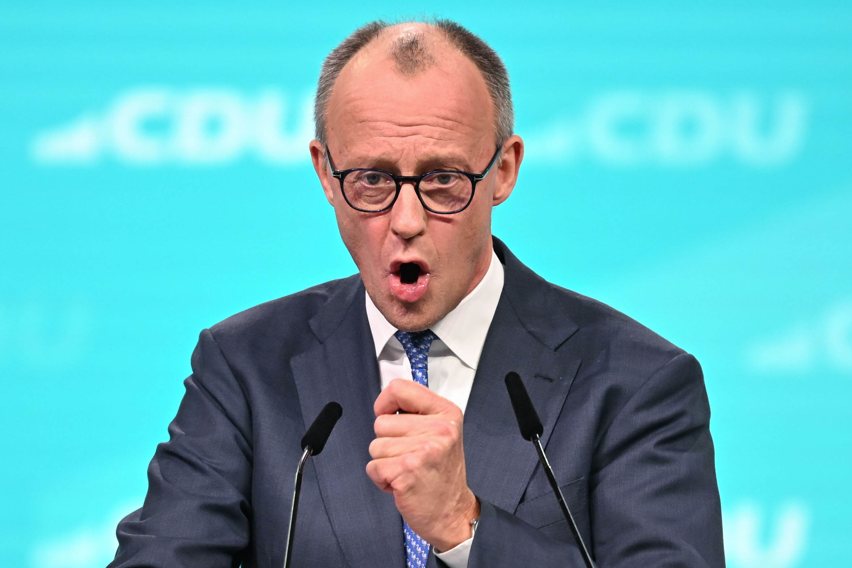 En man i kostym och slips talar vid ett podium med två mikrofoner framför en blå bakgrund med suddig "CDU"-text.