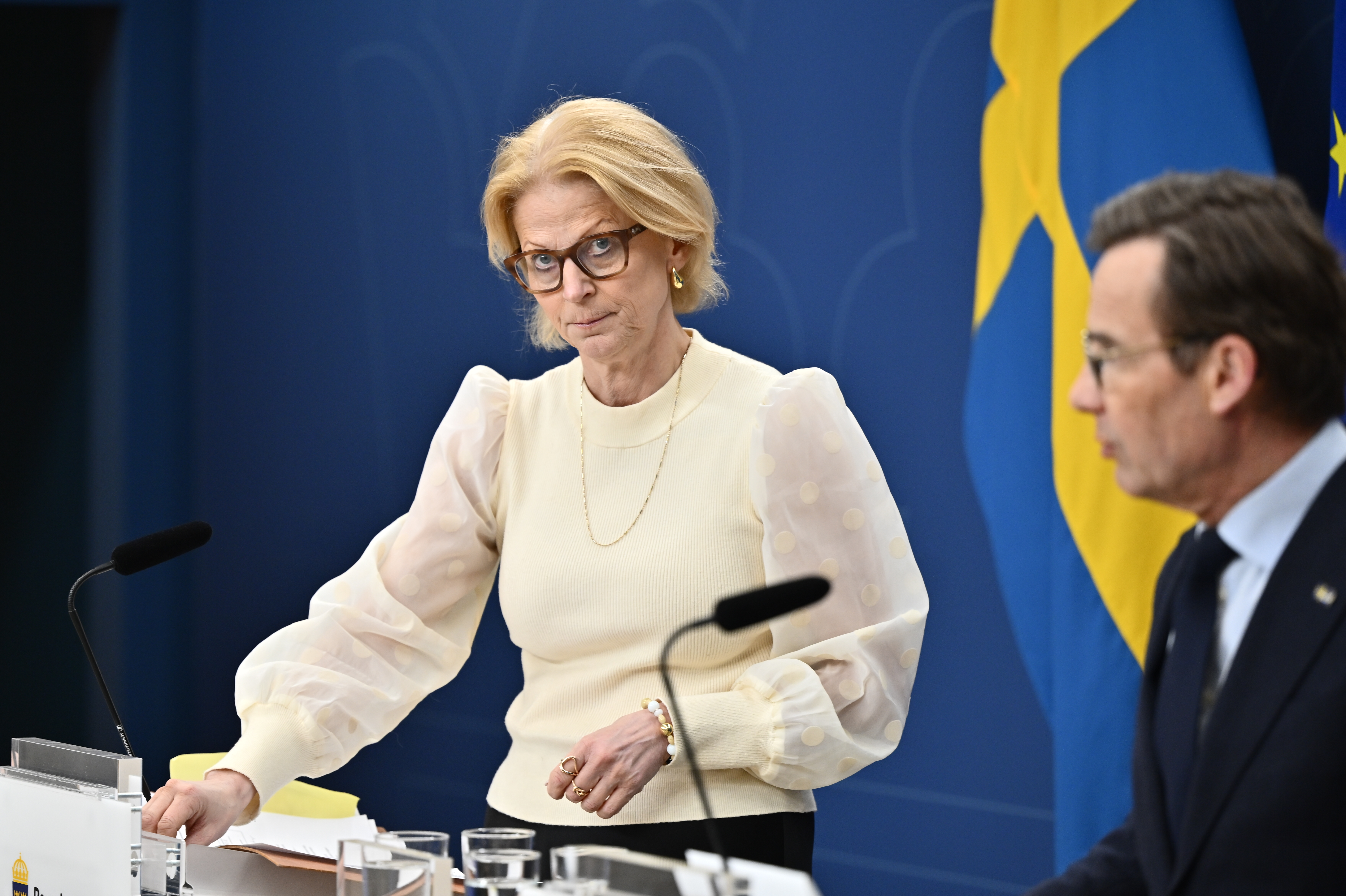 En kvinna i blus står vid ett podium och en man i kostym sitter bredvid henne. En svensk flagga syns i bakgrunden.
