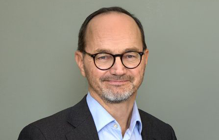 En medelålders man med glasögon, kort hår och skägg har kavaj och ljusblå skjorta, står framför en grå bakgrund.