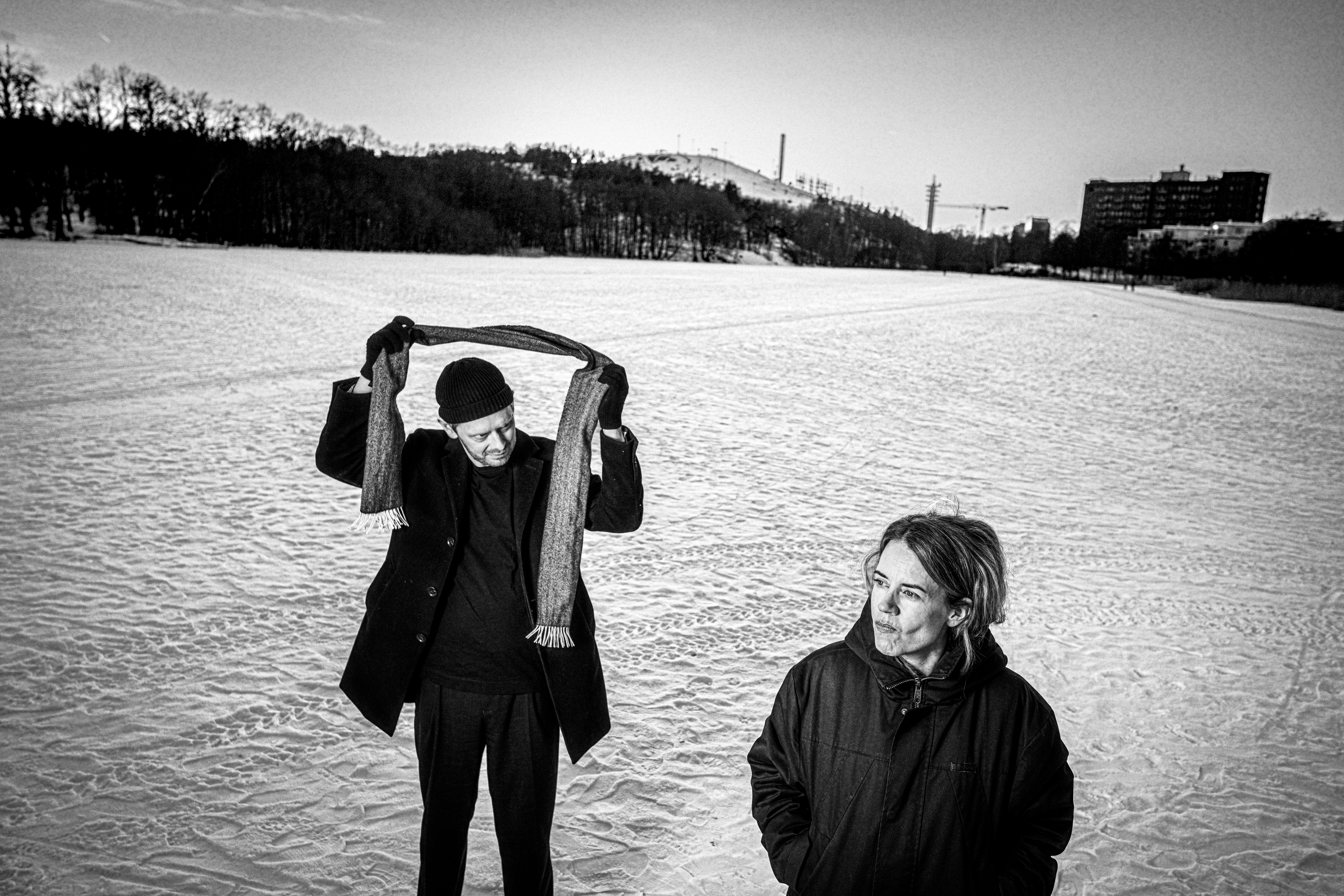Två personer står på ett snötäckt fält; en lyfter en halsduk över huvudet, den andra ser framåt. Träd och byggnader syns i bakgrunden.