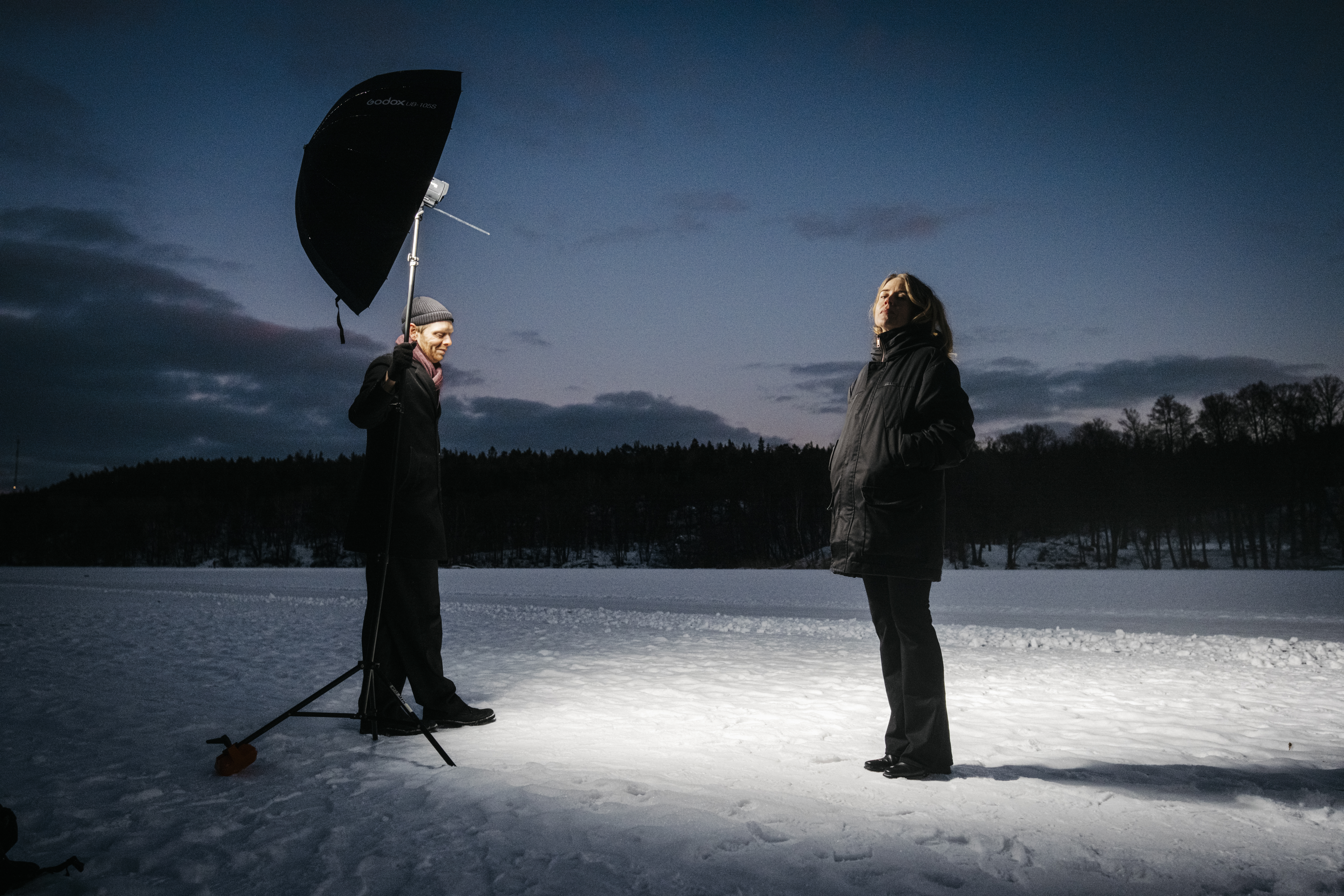 En person med jacka står på snö och lyser upp av en fotolampa som hålls av en annan. Det är skymning. Träd och moln syns bakom.