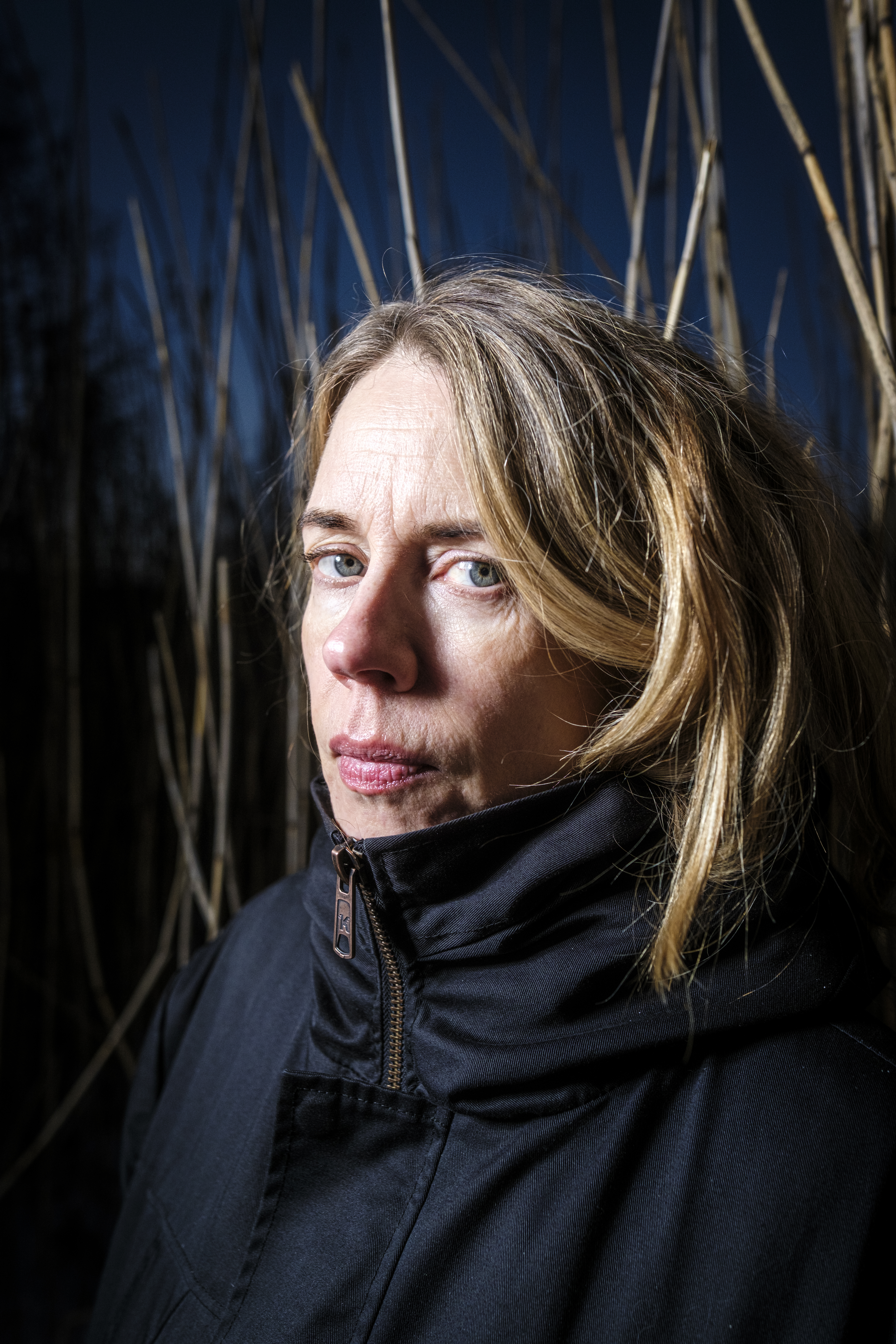En kvinna med blont hår i svart kappa står utomhus framför höga smala grenar på natten och ser rakt mot kameran med allvarlig min.