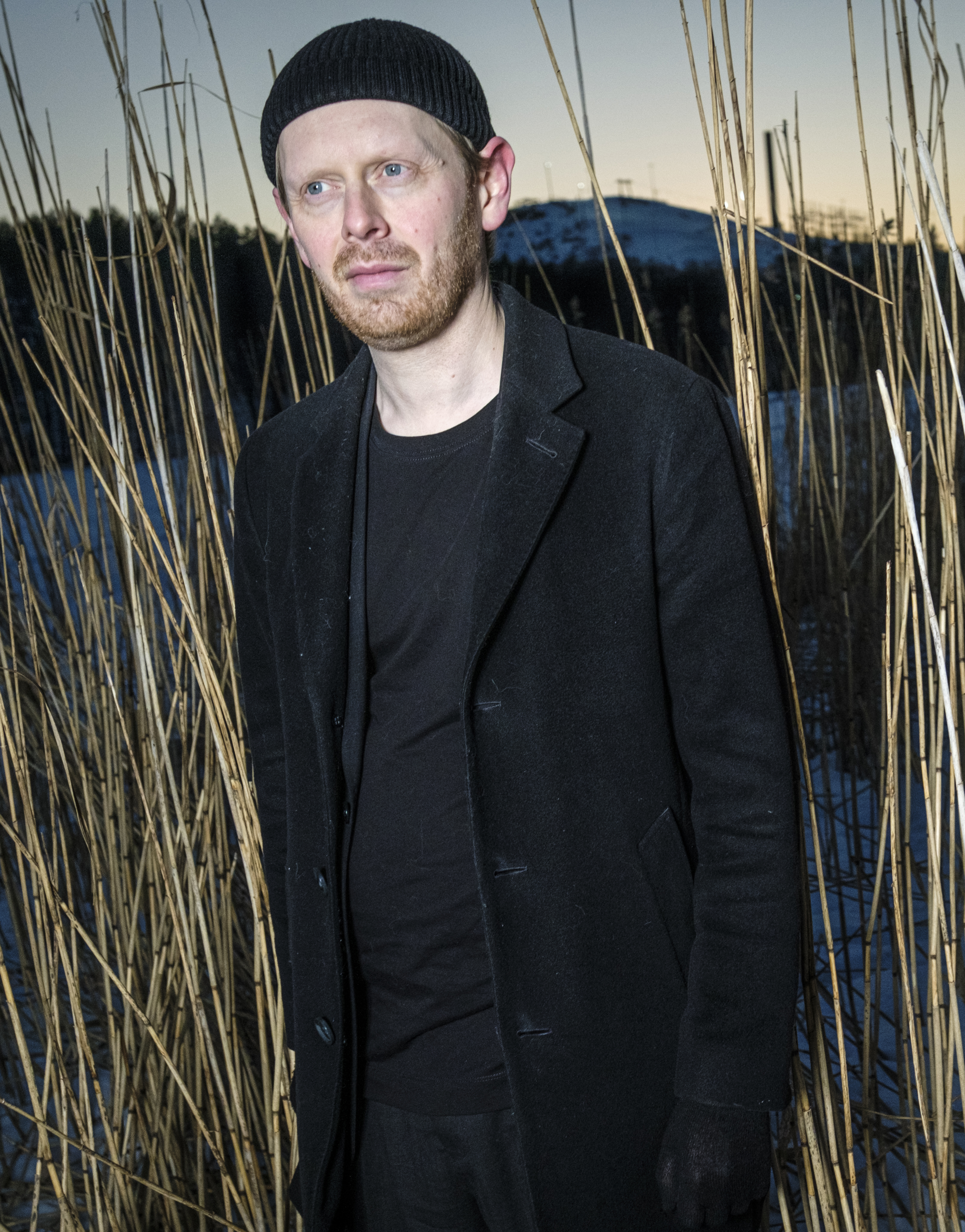 En man med svart rock och mössa står bland höga vass utomhus i skymningen, med snö på marken och en kulle bakom.