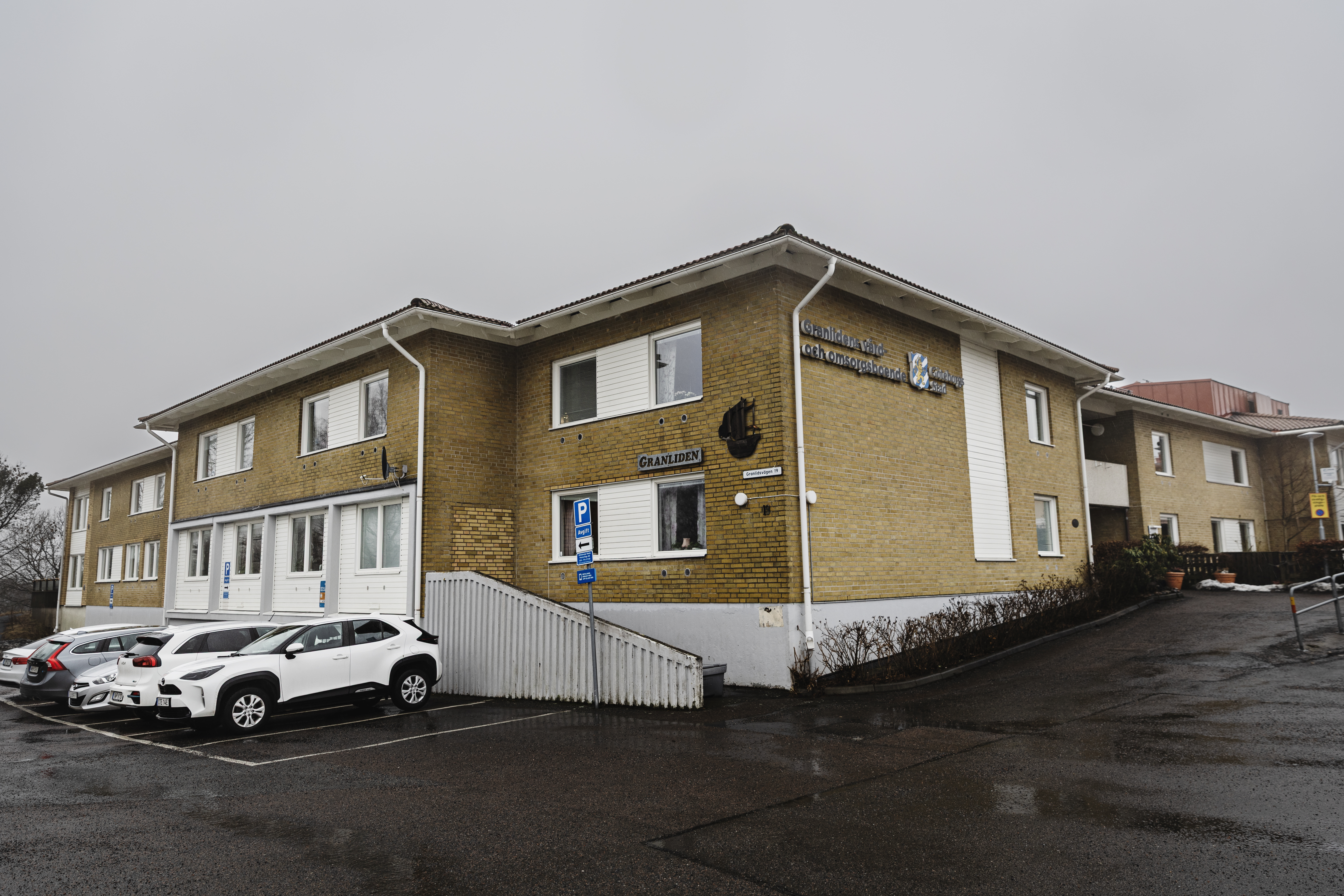 En tvåvånings byggnad i gul tegel med vit list, ramp för tillgänglighet, parkerade bilar framför och skyltar om verksamheter.