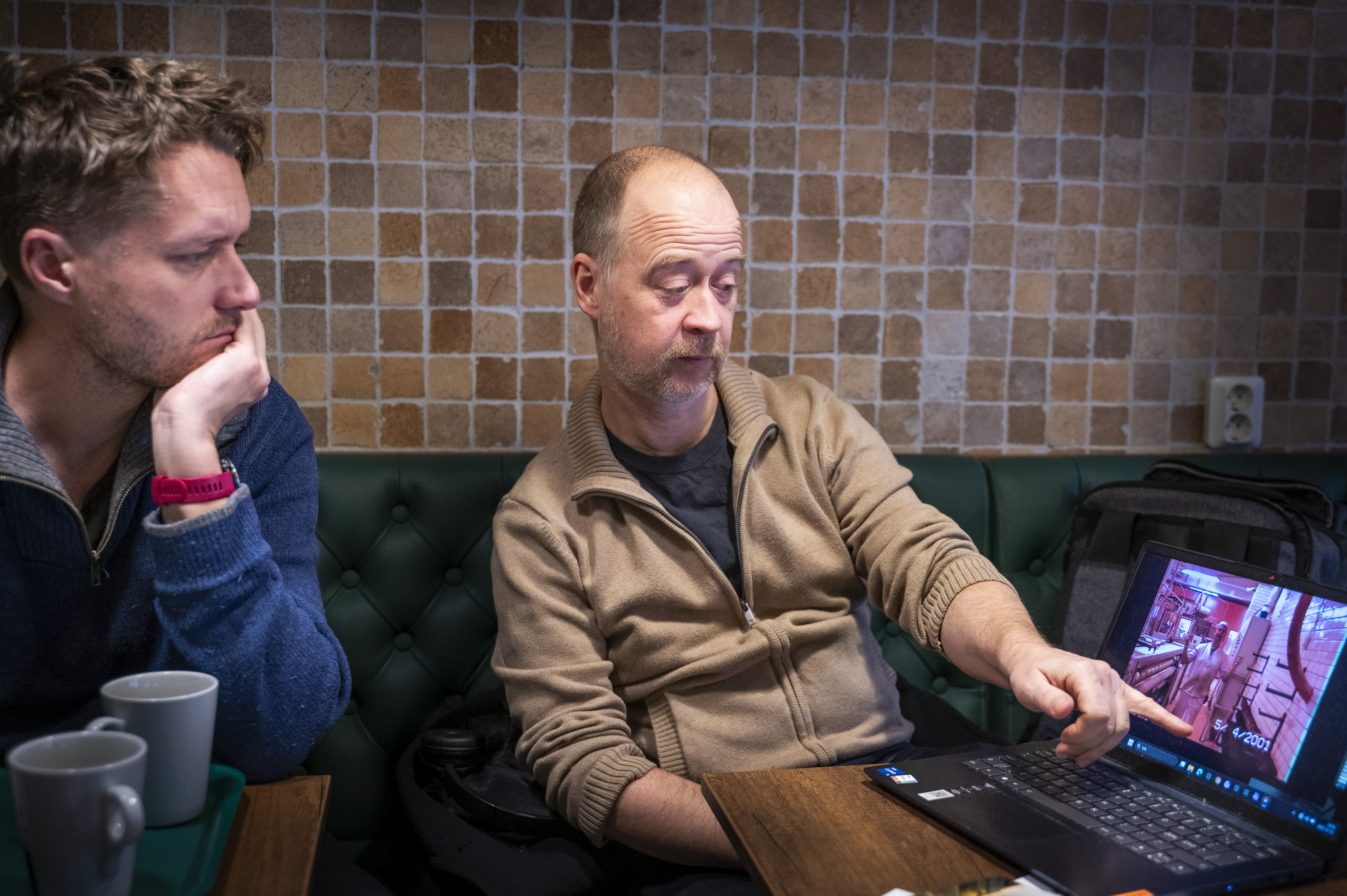 Två män sitter vid ett bord med kaffemuggar, en pekar på något på en bärbar dator som visar filmklipp från ett bageri.