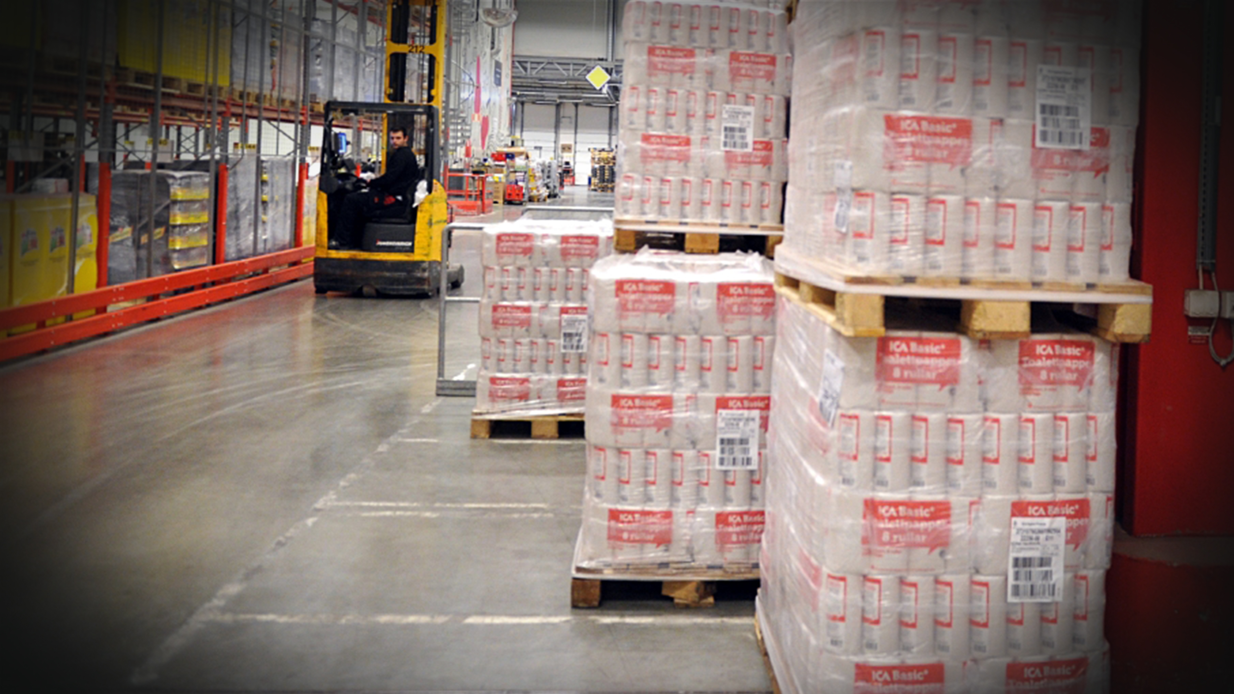 Ett lager med staplade pallar inslagna i plast och en arbetare kör truck i bakgrunden.