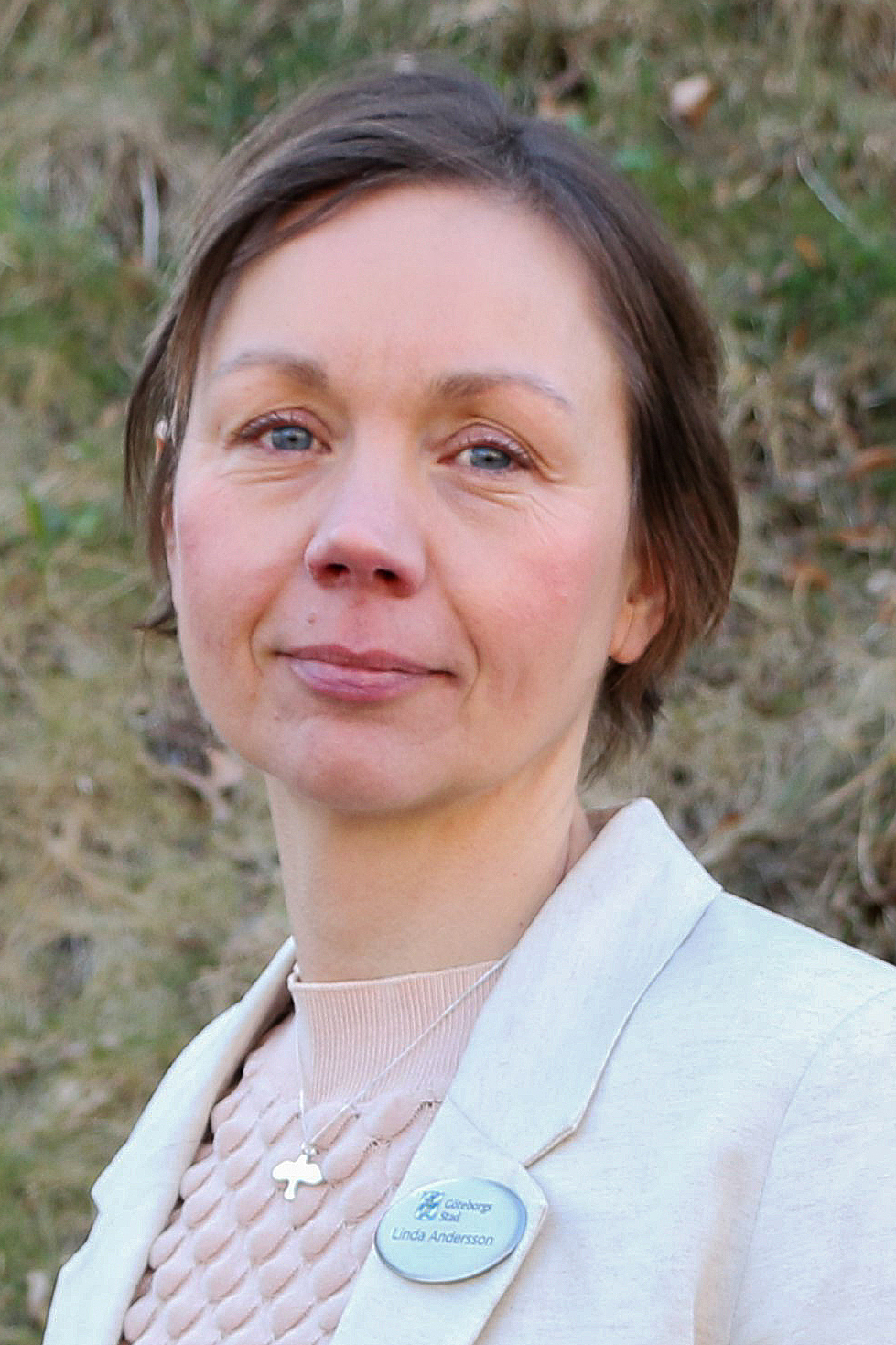 Linda Andersson med kort brunt hår, ljus kavaj och namnbricka står utomhus framför en gräsyta.