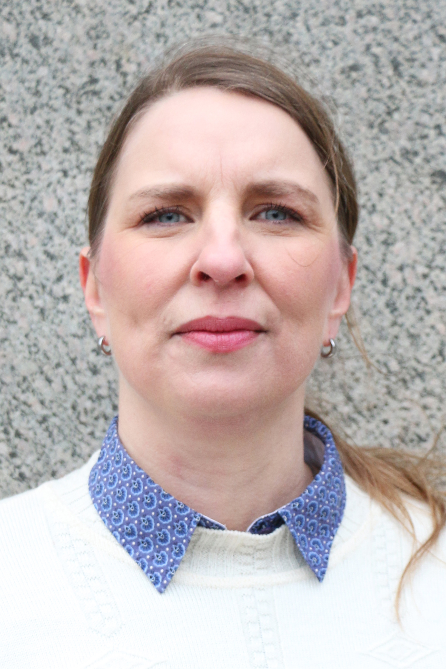 Sophia Furström, med ljus hy, blå ögon och ljusbrunt hår, stirrar på kameran framför en stenmur. Hon är klädd i skjorta och tröja.