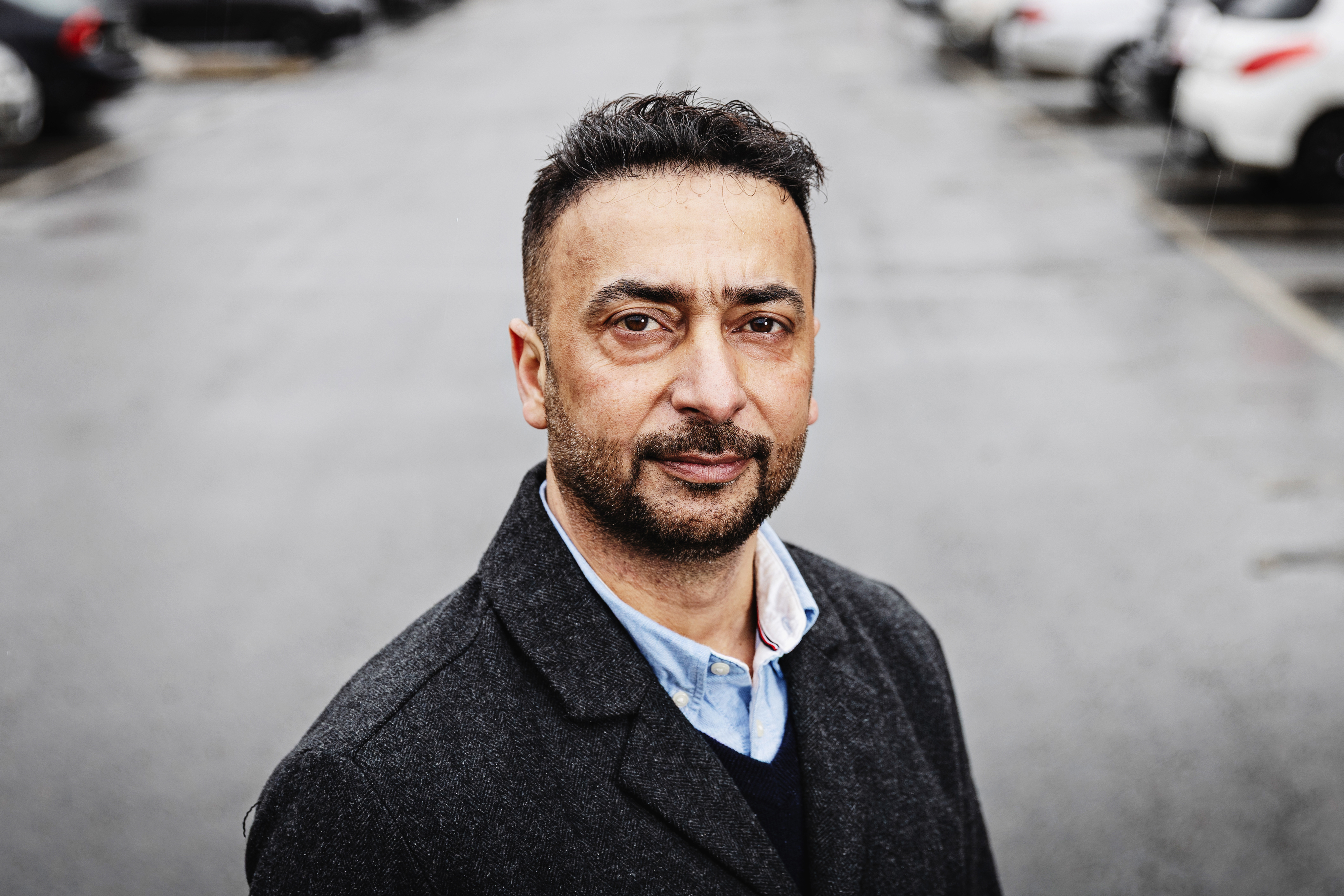 Rabie al-Rubeai, med kort svart hår och skägg, står på en tom parkeringsplats under en molnig himmel, iförd en svart kappa över skjortan.