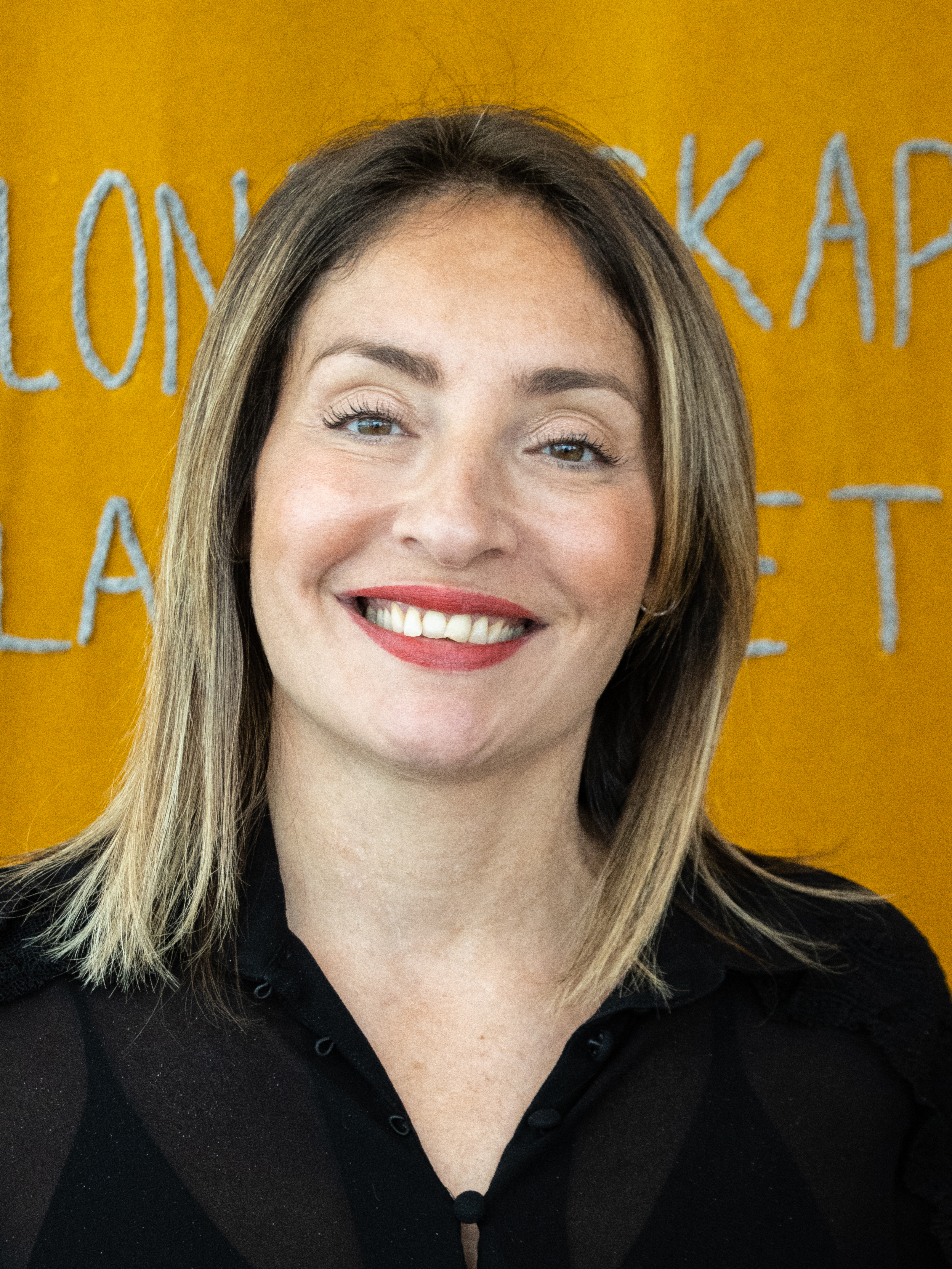 Deanna Hylan, med axellångt rakt blont hår och iklädd en svart topp, ler framför en gul bakgrund med delvis synlig vit text.