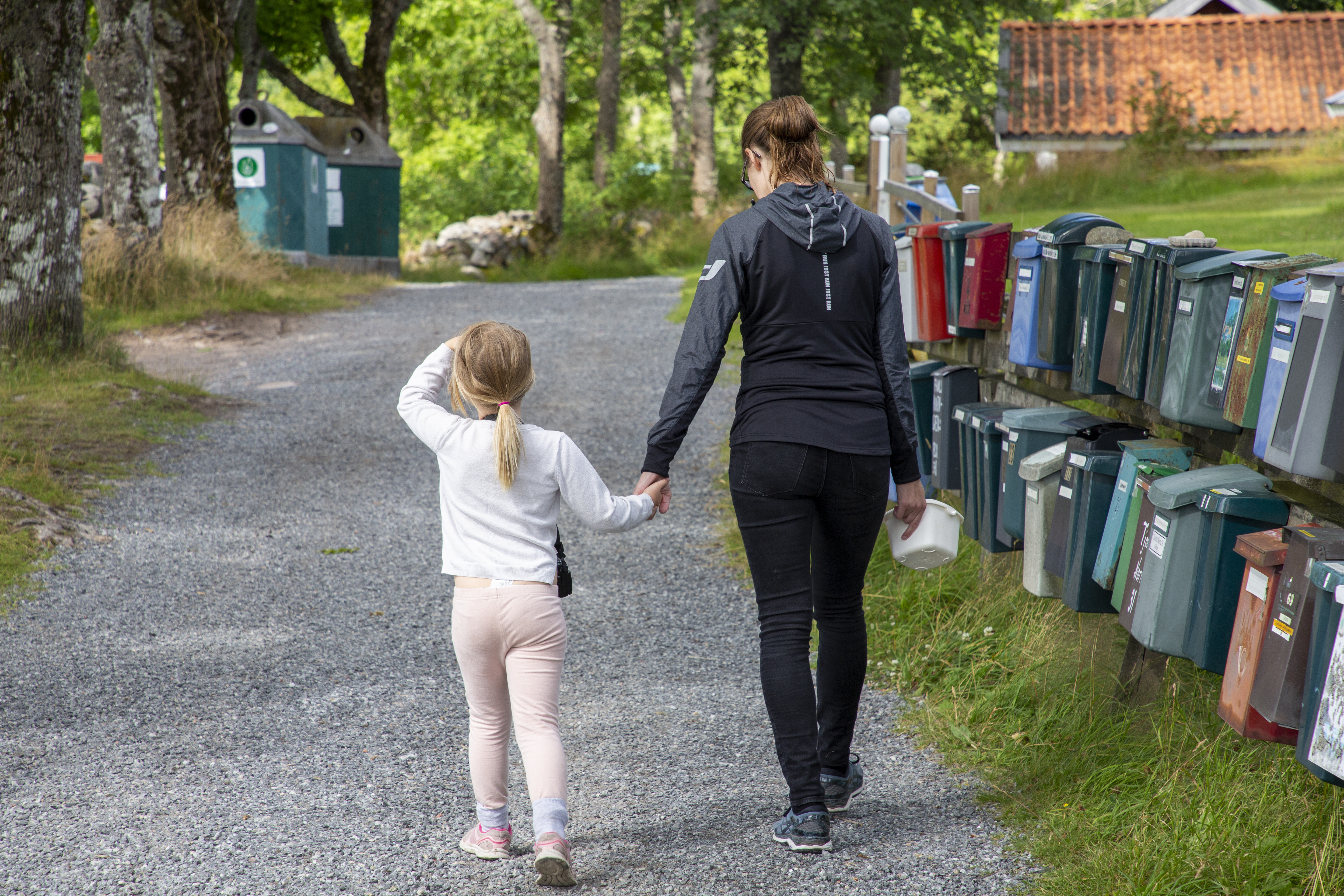 En vuxen och ett barn går hand i hand på en grusväg med brevlådor, omgivna av träd och växter.