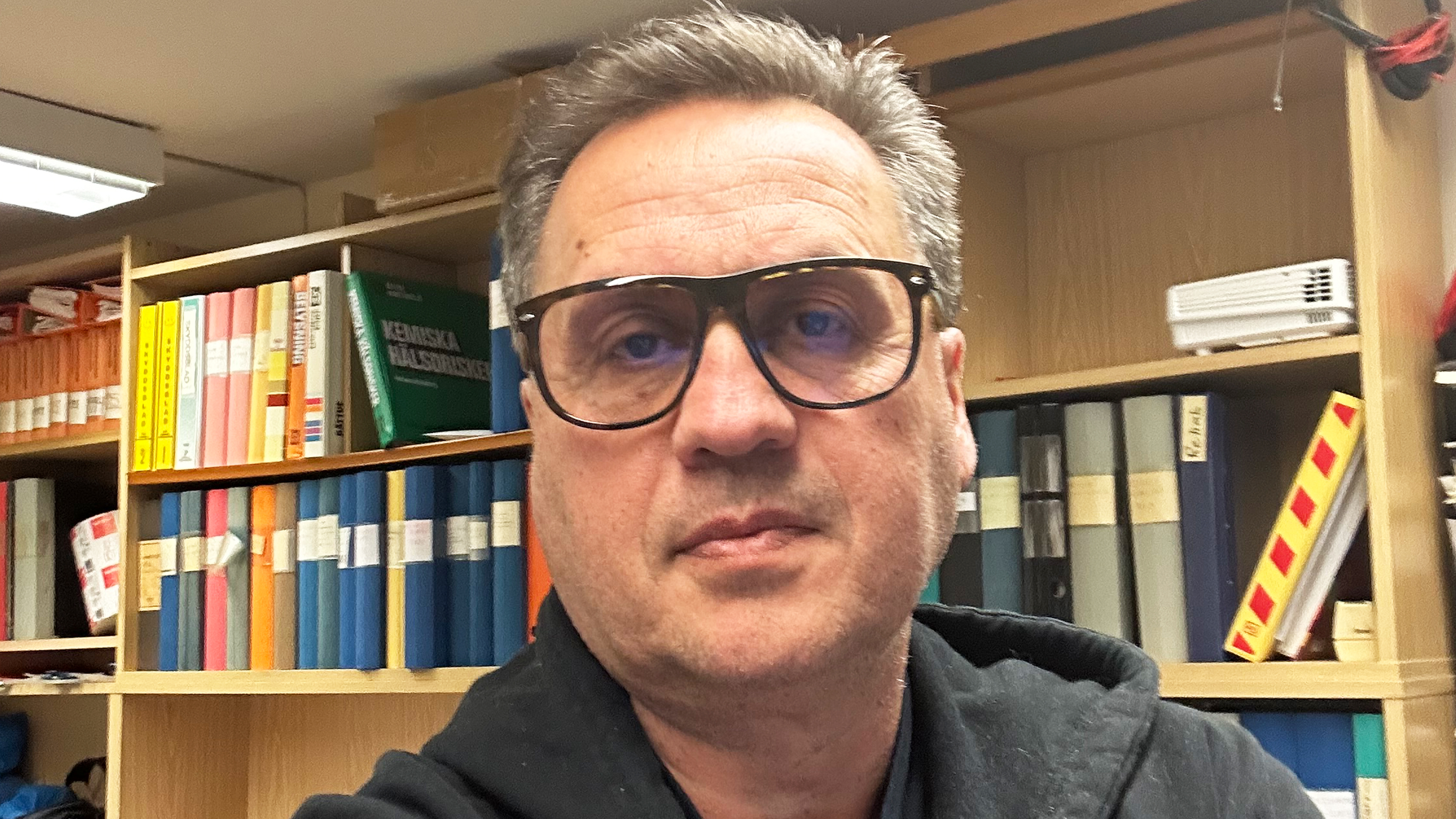 En man med glasögon och mörk skjorta sitter framför hyllor med böcker, pärmar och kontorsmaterial.