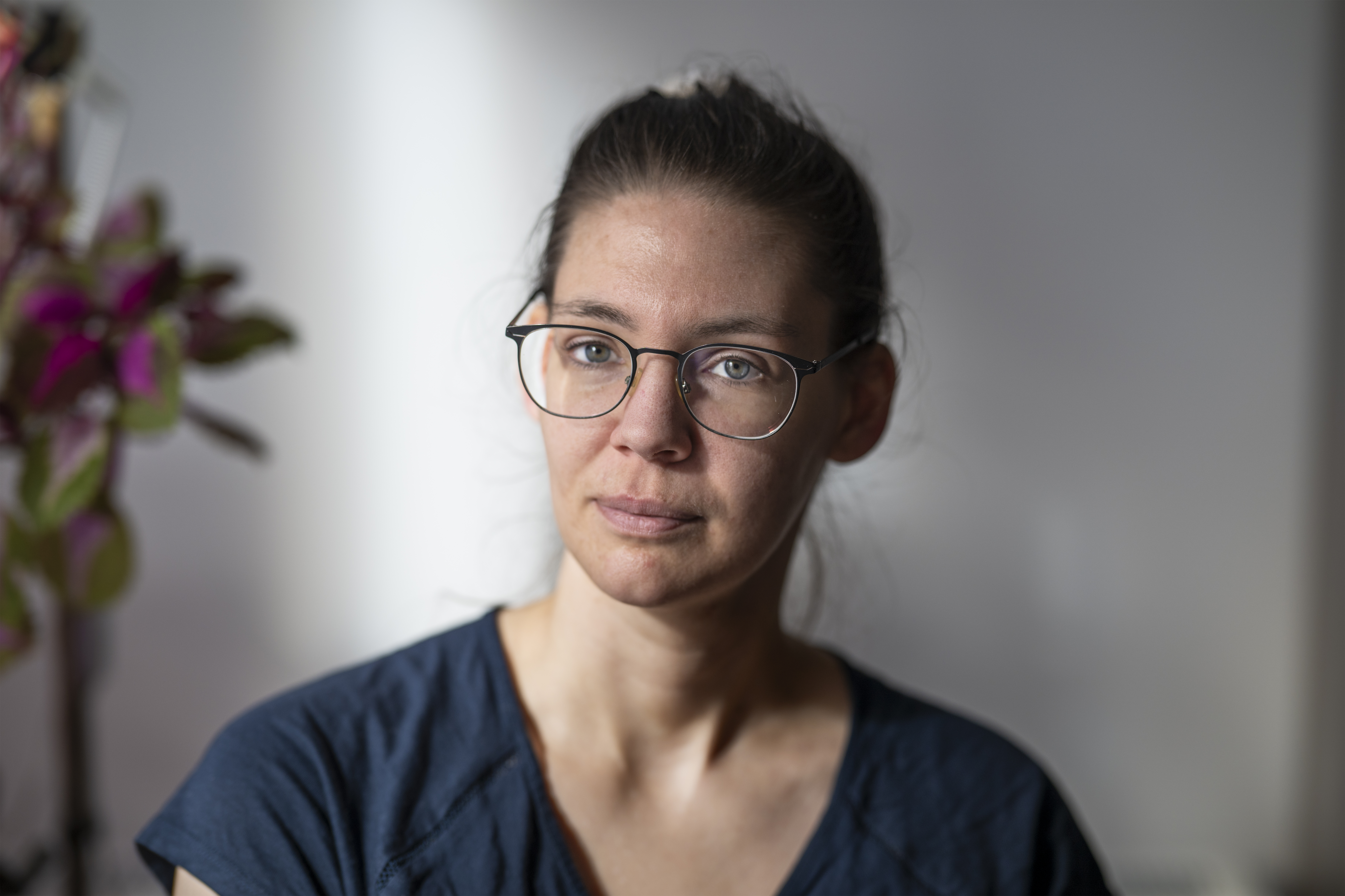 Sara Eberhag med glasögon och brunt hår uppsatt tittar mot kameran och har en mörkblå topp. En växt syns i bakgrunden.
