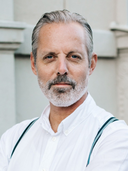 Dan-Ove Marcelinde, gråhårig och skäggig, iklädd vit skjorta och hängslen, står utanför framför en starkt upplyst byggnad.