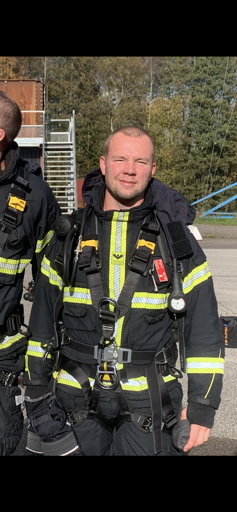 En brandman i full utrustning står utomhus, med träd och träningsutrustning i bakgrunden.