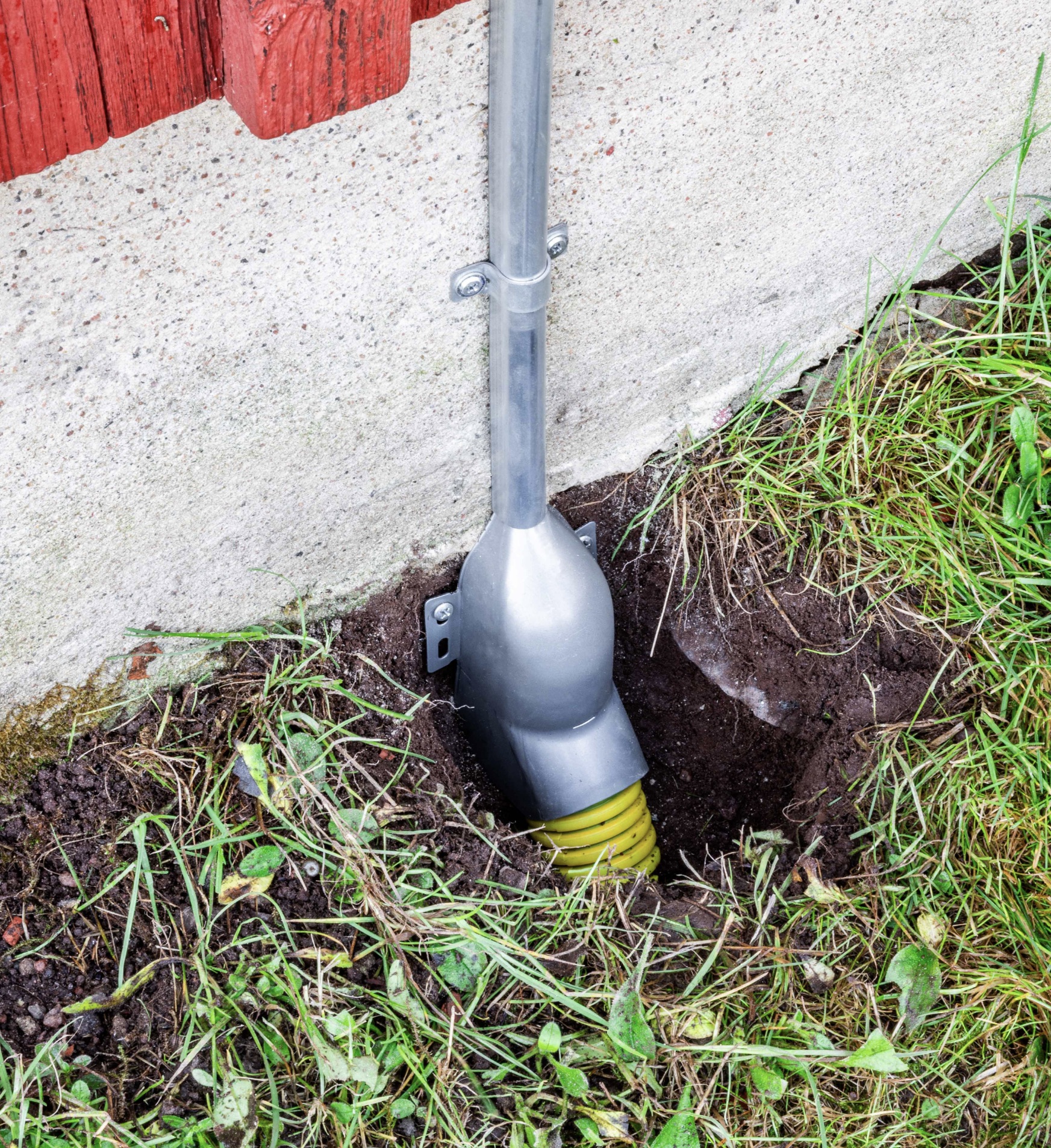 En stupränna i metall med en gul korrugerad dräneringsslang är installerad i ett hål bredvid en husgrund och gräs.