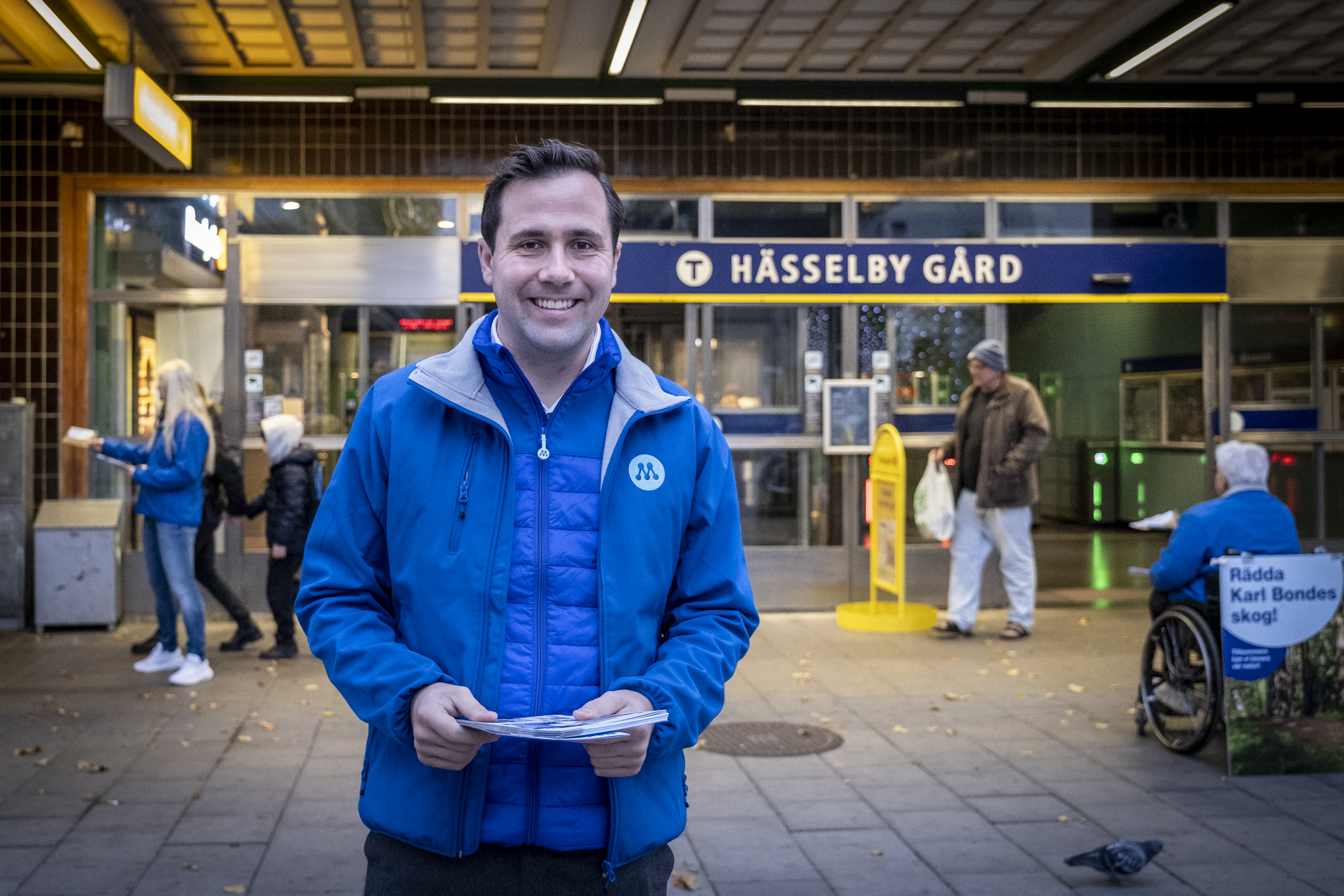 En man i blå jacka står utanför Hässelby Gård tunnelbanestation och håller papper. Flera personer syns i bakgrunden.