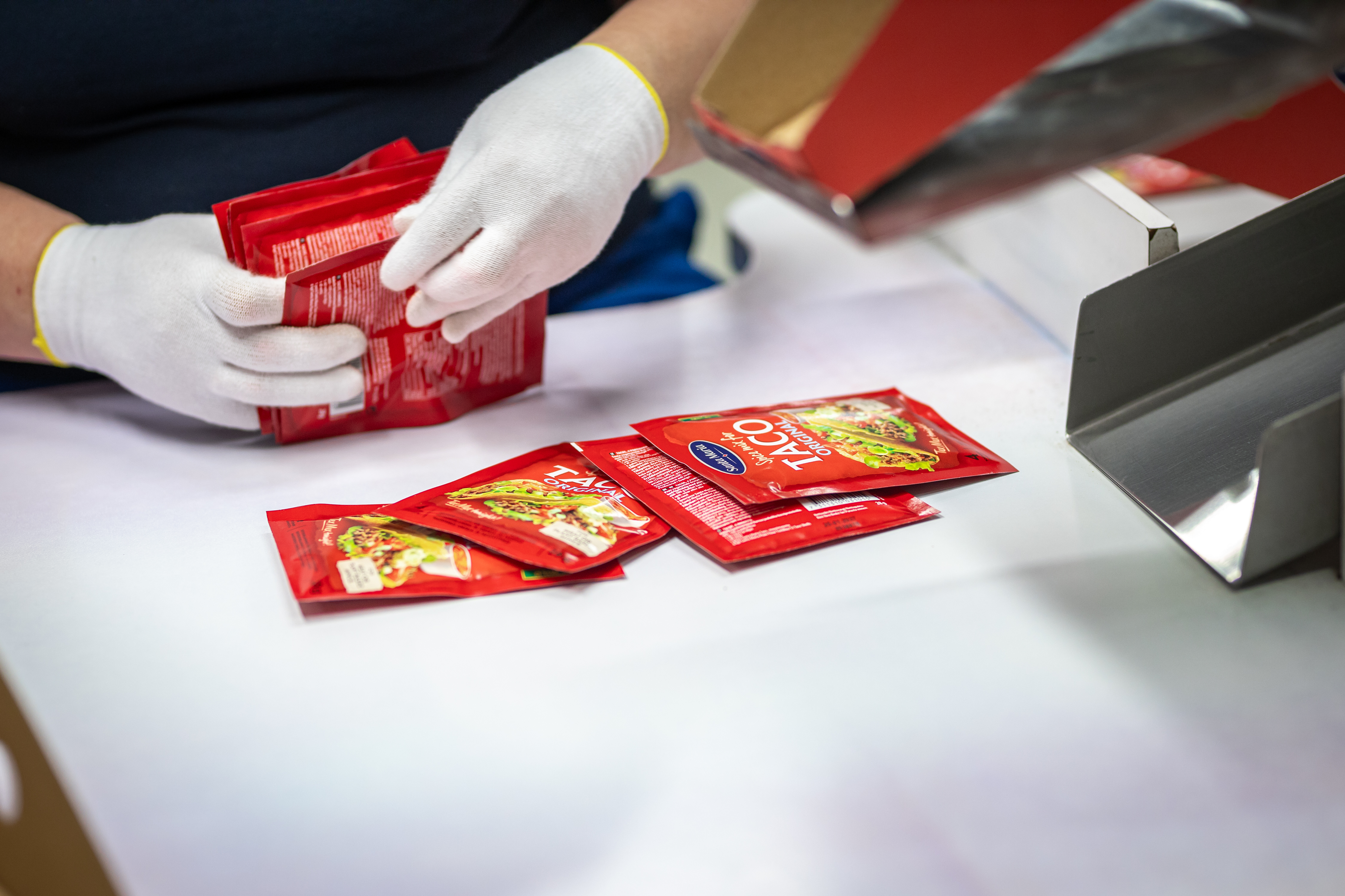 En person med vita handskar hanterararar flera röda förpackningar märkta "Taco Seasoning" på en vit yta nära metallutrustning.
