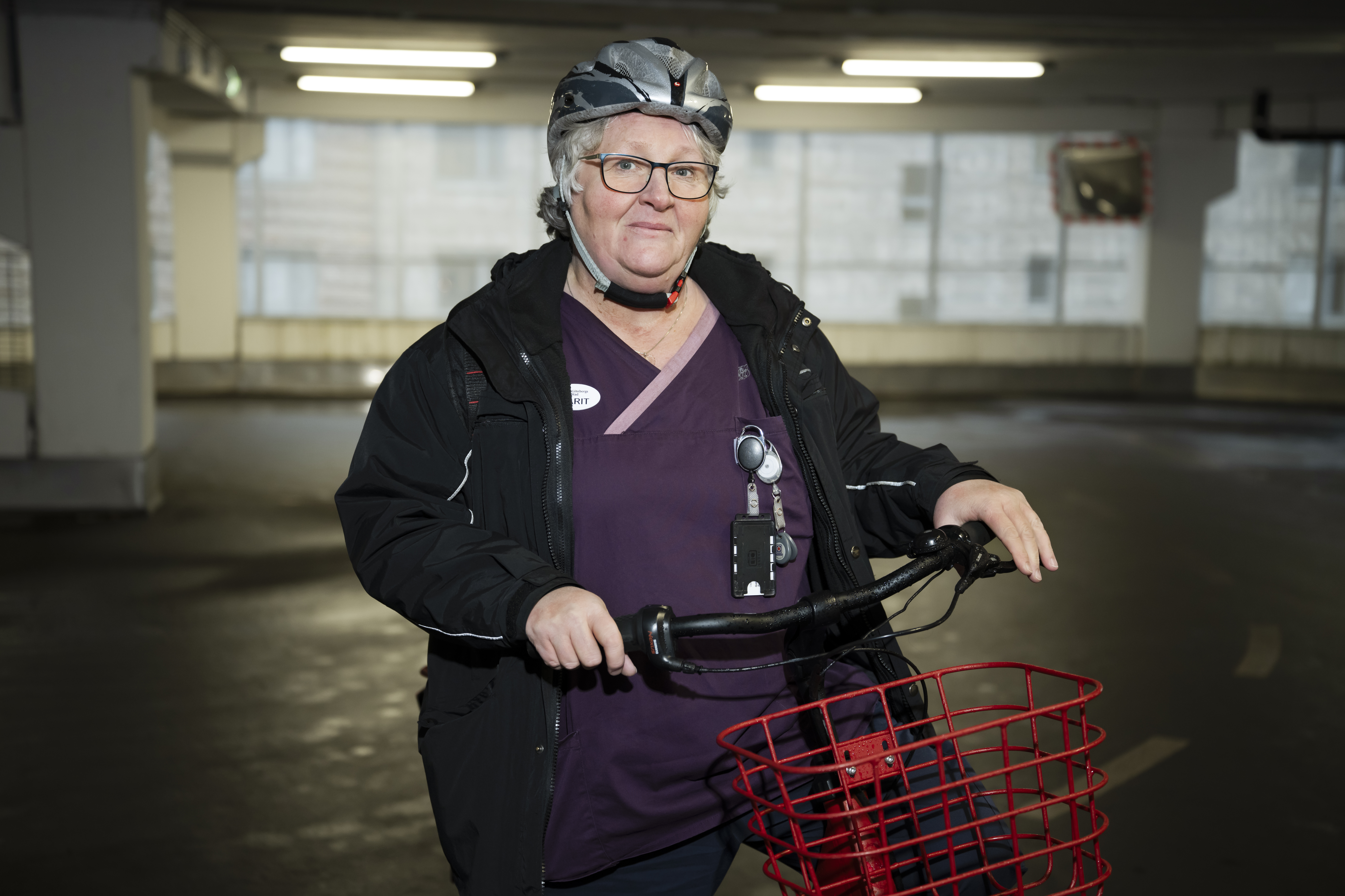 En person med hjälm och arbetskläder står inomhus och håller i en cykel med röd korg i ett parkeringsgarage.