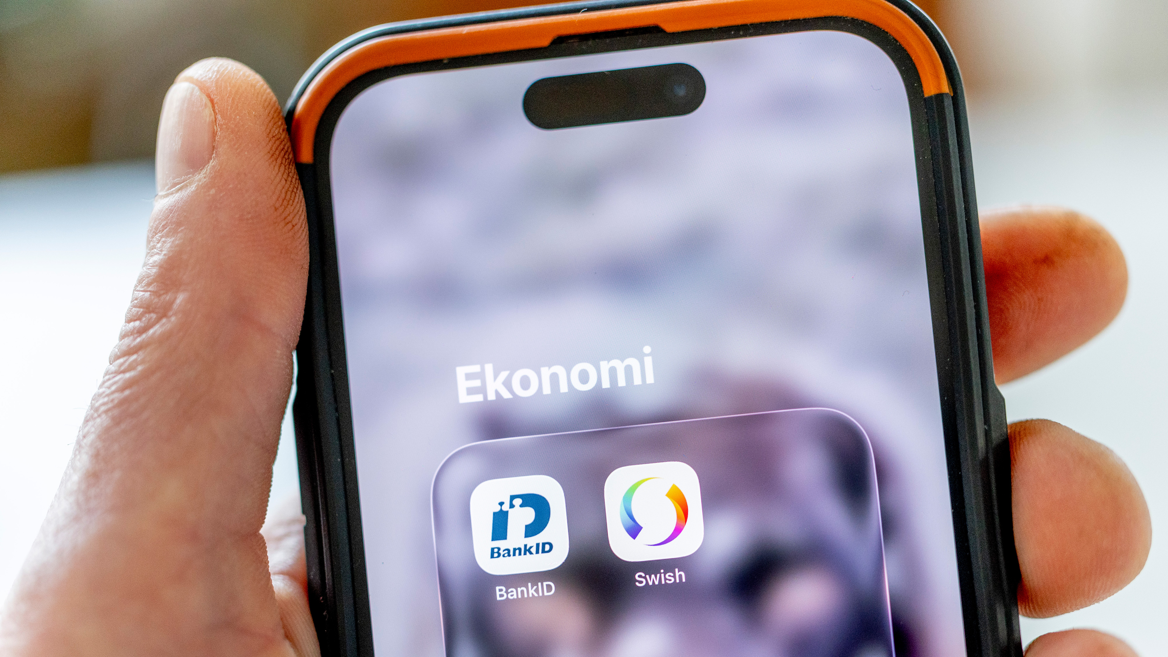 En hand håller en mobiltelefon som visar en mapp med texten "Ekonomi" och ikoner för BankID och Swish på skärmen.