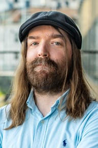 En person med långt brunt hår, helskägg och mustasch och ljusblå skjorta står utomhus i stadsmiljö.