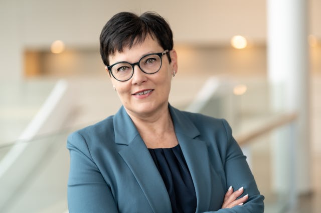 En kvinna med kort mörkt hår, glasögon och en blå kavaj står inomhus med armarna i kors.