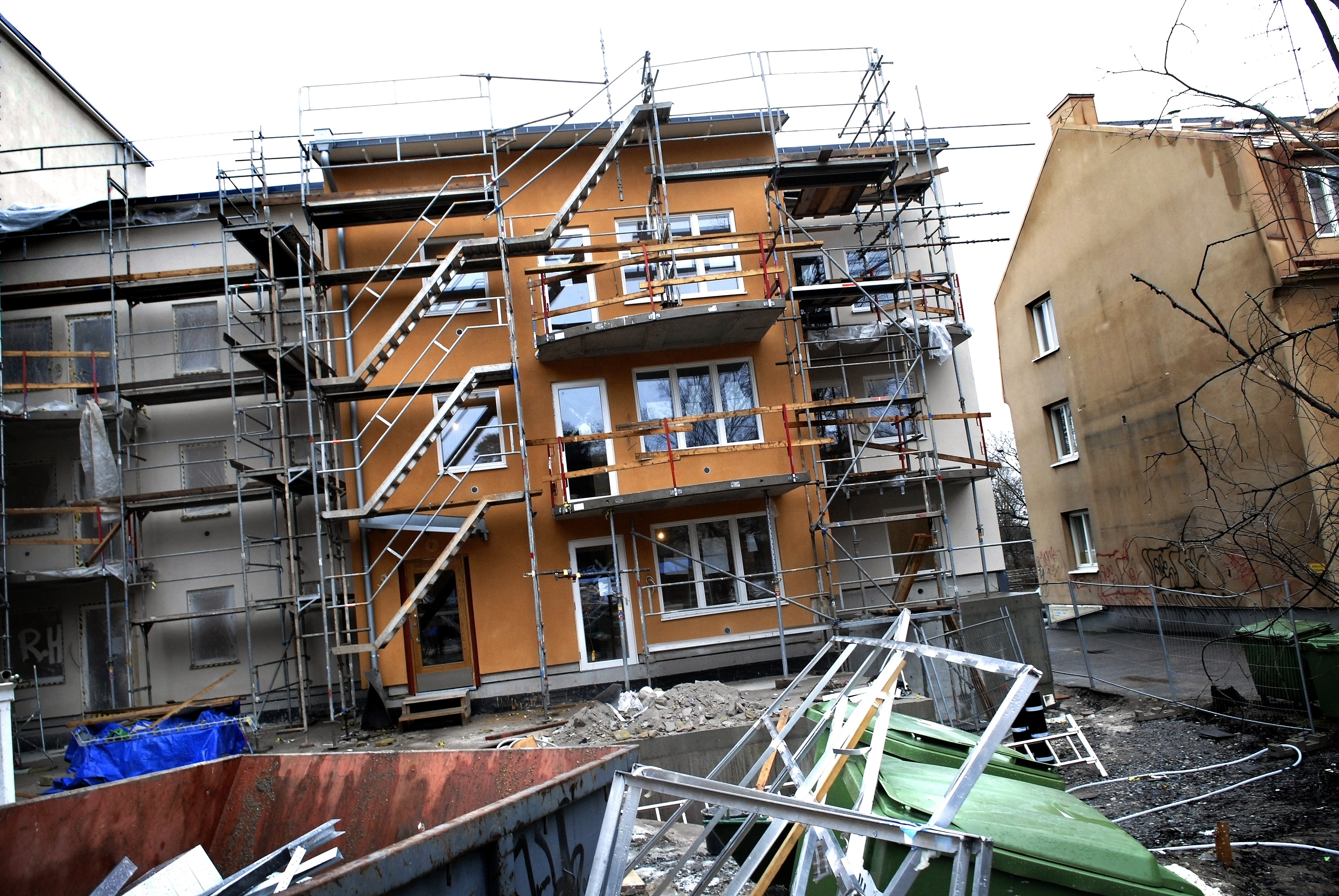 En orange byggnad under uppförande med byggställningar, omgiven av bråte, byggmaterial och containrar i förgrunden.