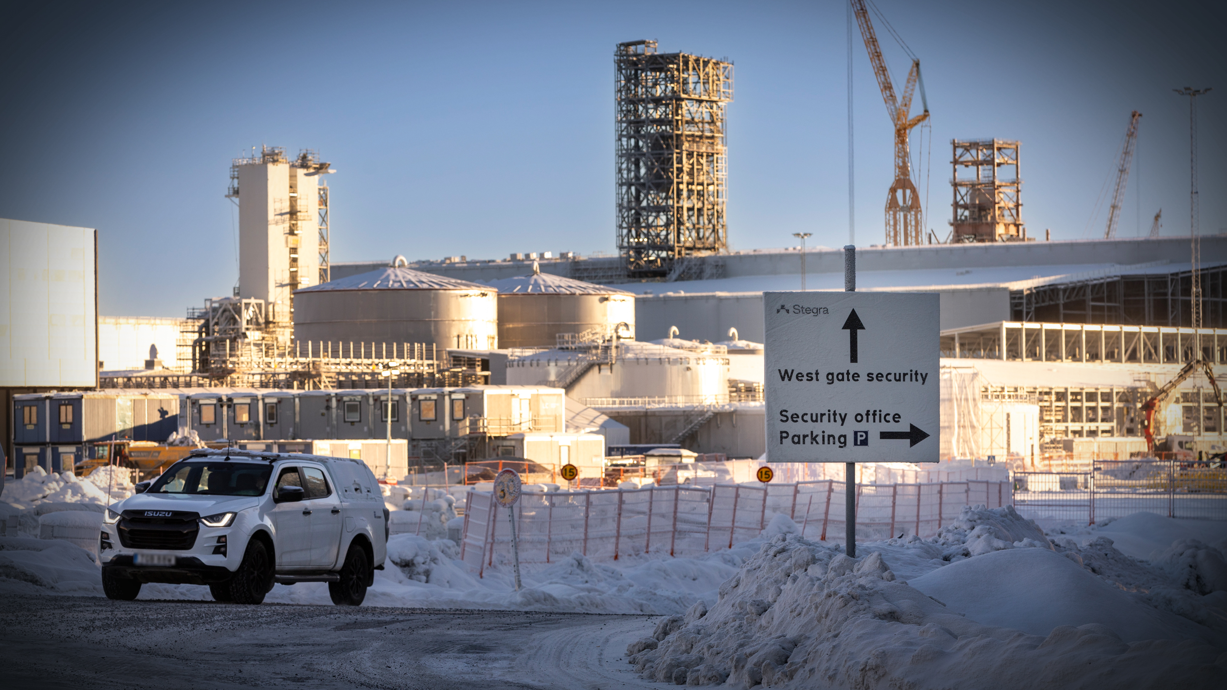 En vit SUV kör på en snötäckt väg nära en industribyggnad med byggkranar och säkerhetsskyltar i förgrunden.