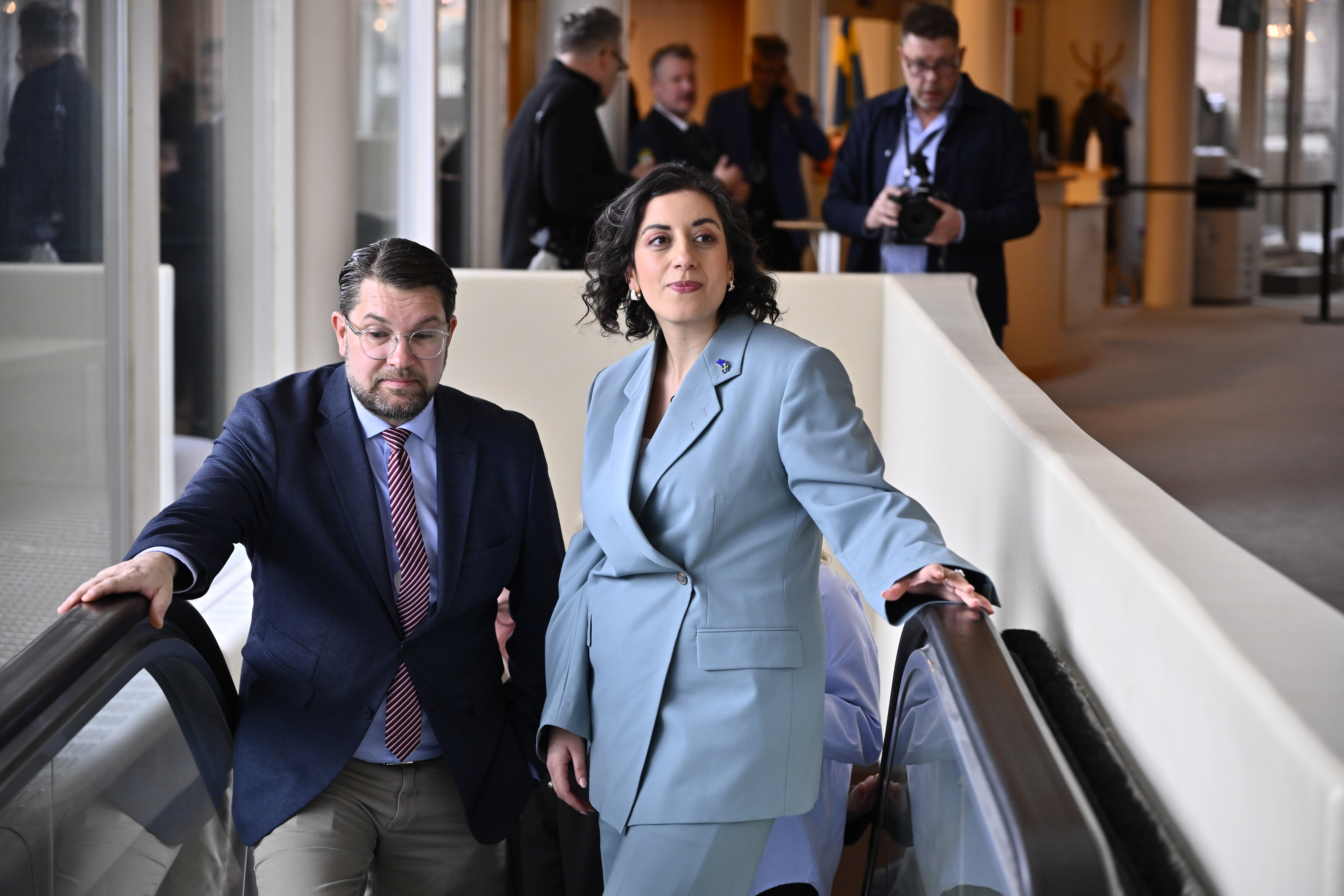 Två personer i affärskläder åker rulltrappa, flera andra står i bakgrunden, några håller kameror, inomhus.