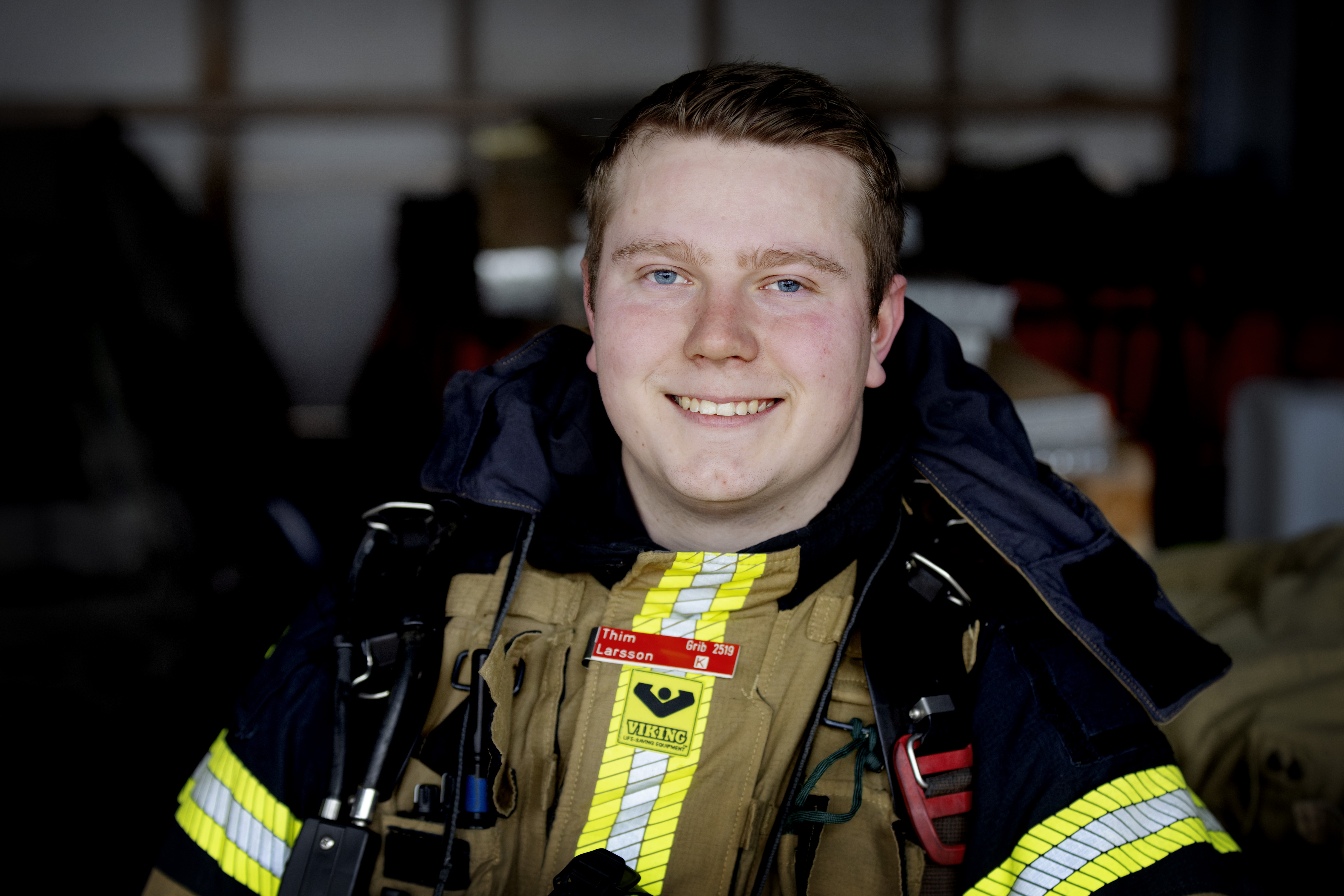 En brandman i uniform tittar mot kameran och står inomhus med räddningsutrustning i bakgrunden.