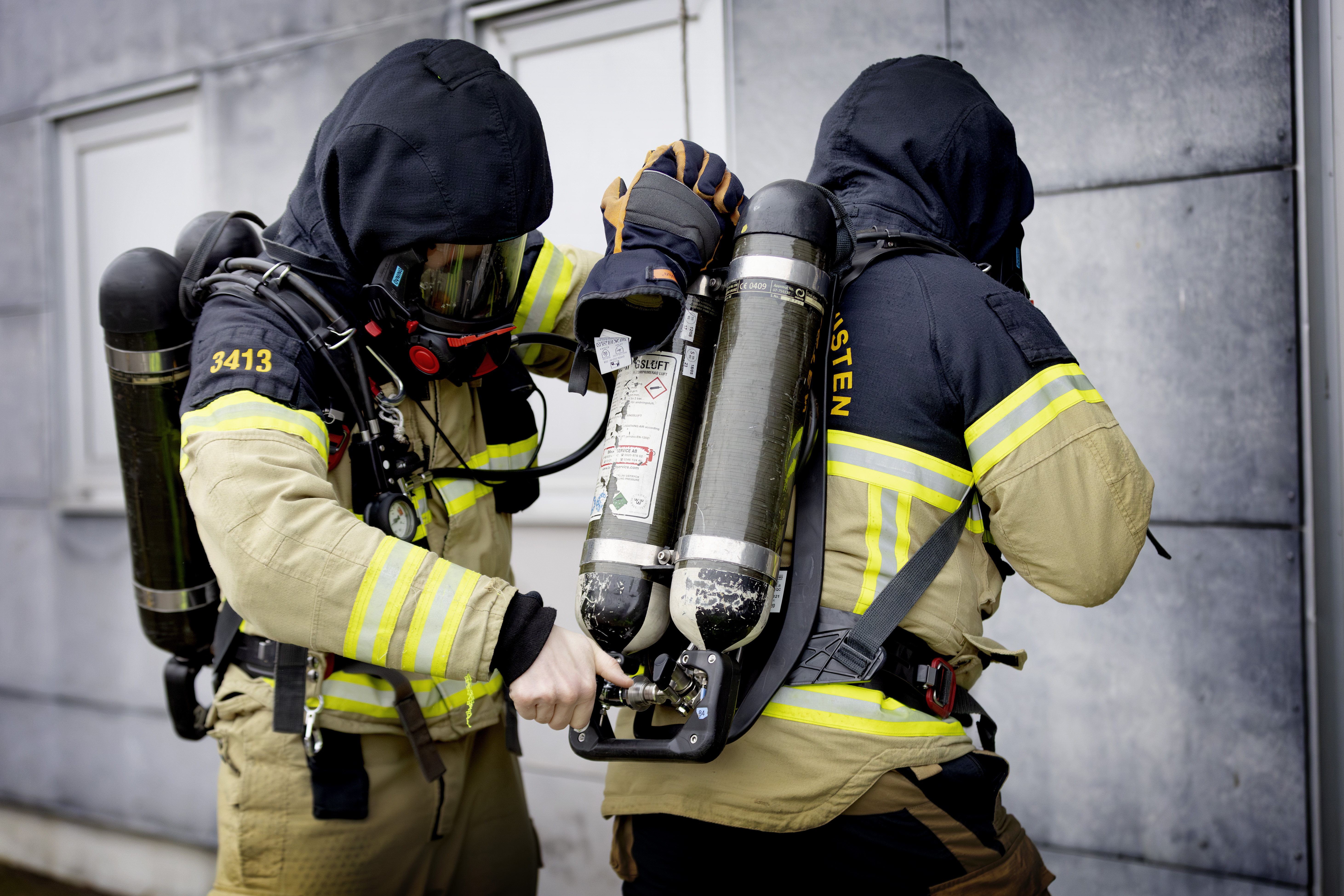 Två brandmän i arbetskläder justerar varandras syrgastuber inför att de ska gå in i en byggnad.