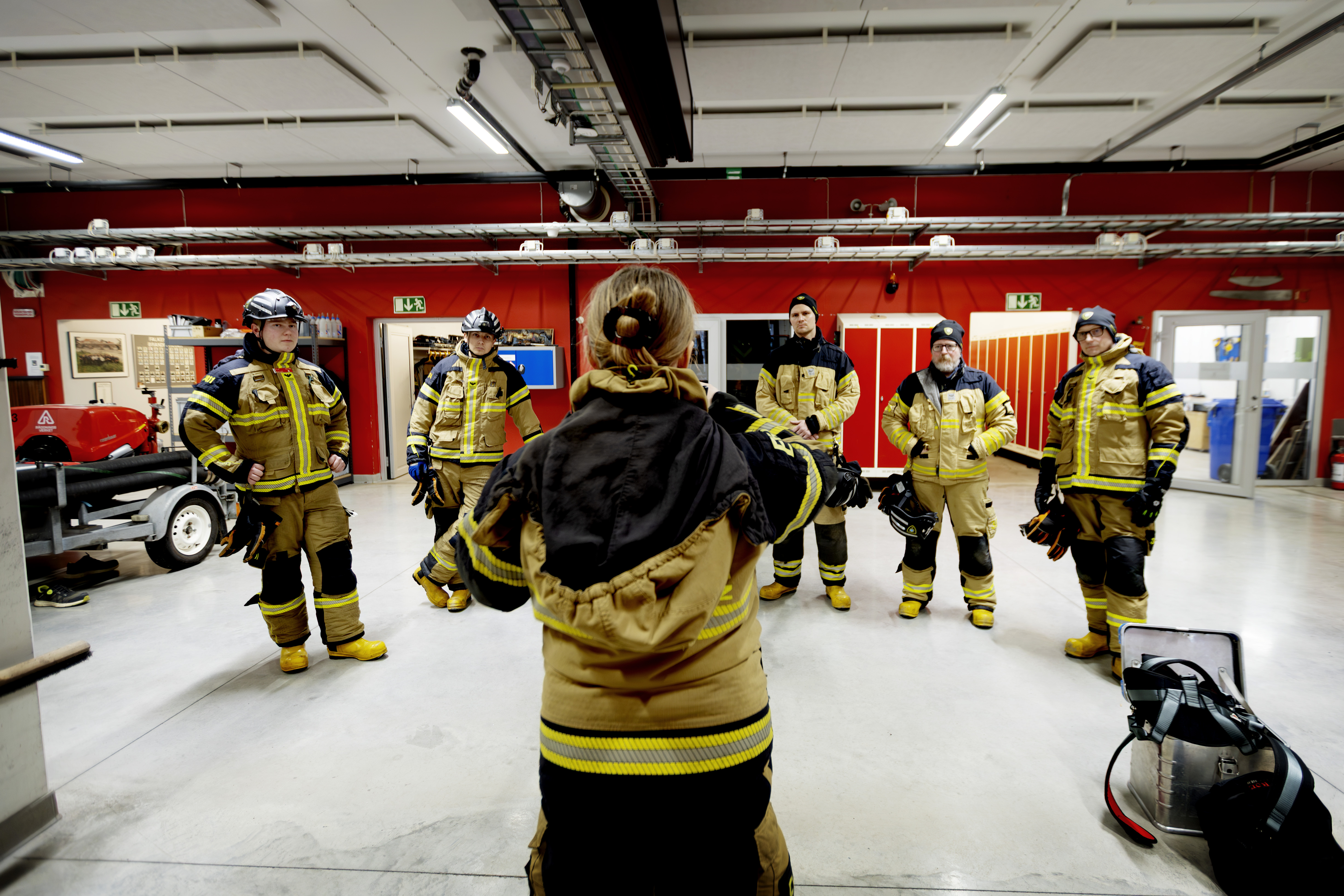 En grupp brandmän i utrustning står i en brandstation och lyssnar på en person som ger instruktioner.