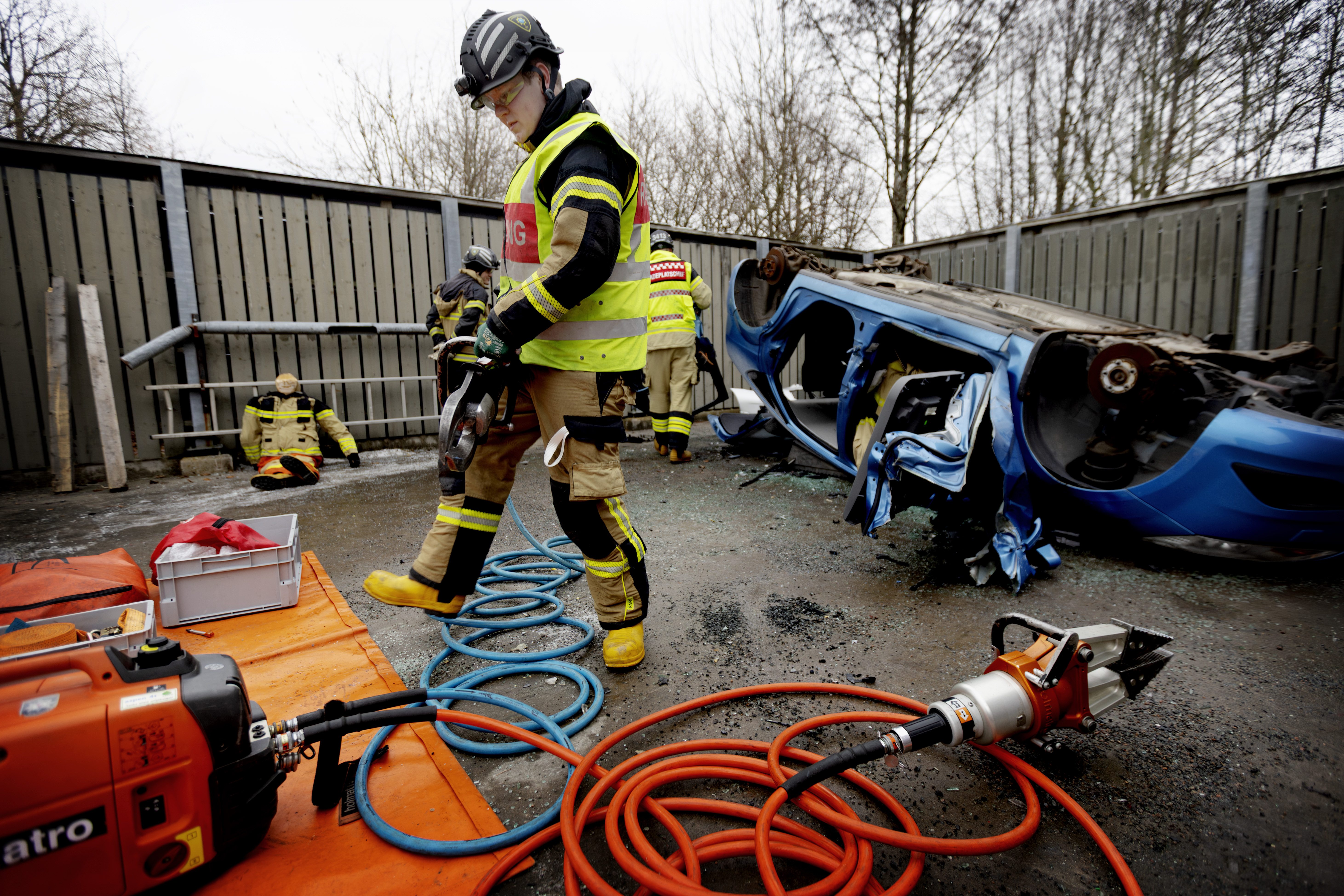 Brandmän med skyddsutrustning övar räddning med hydraulverktyg vid en vält och skadad blå bil på en utomhusövningsplats.