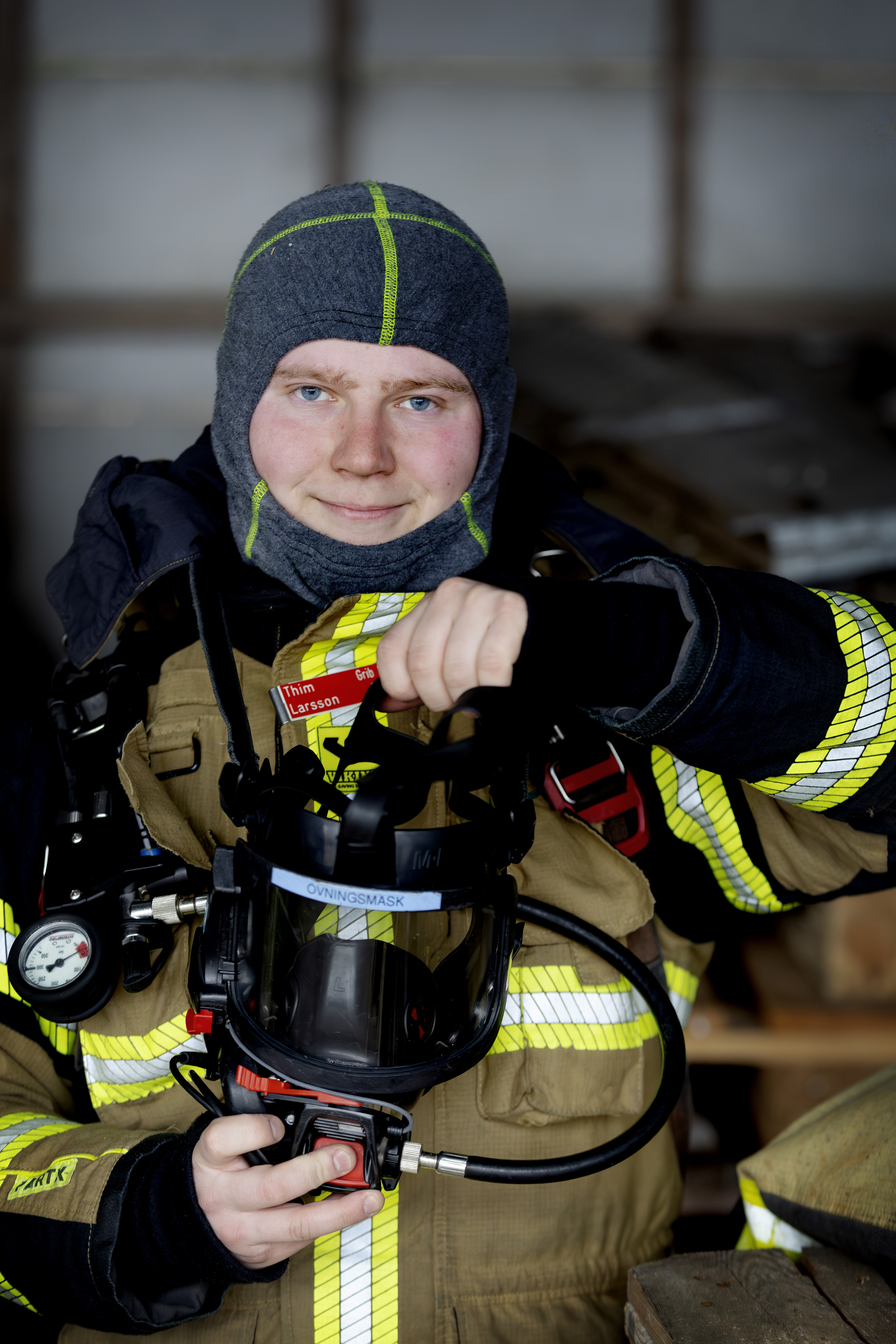 En brandman i skyddsutrustning håller i en andningsmask och står inomhus med utrustning synlig i bakgrunden.