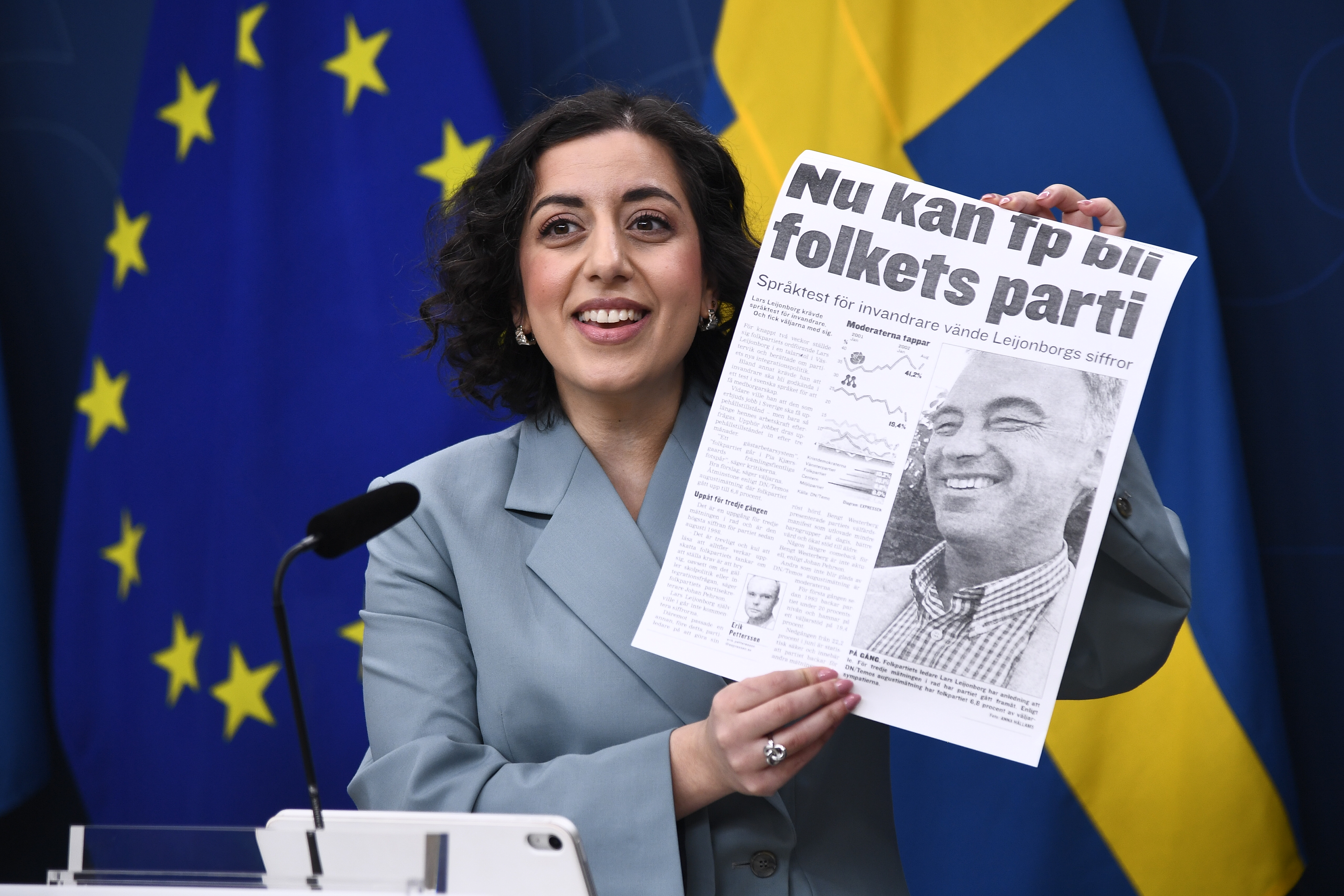 En kvinna i kostym håller upp en tidningsartikel med rubrik och foto, framför EU- och svenska flaggor.