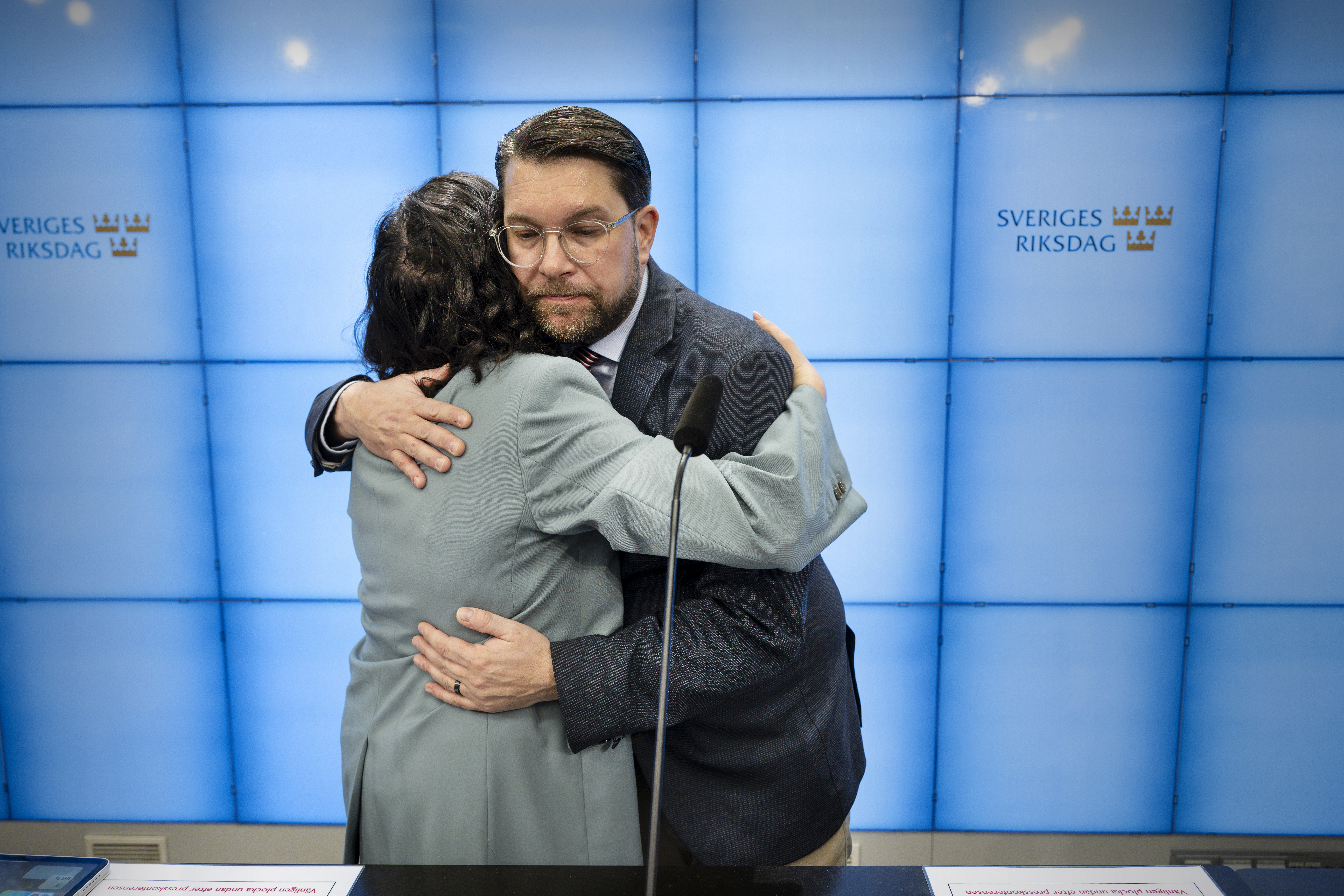 Två personer kramas framför en blå skärm med Sveriges riksdags logotyp och står bakom mikrofoner på en presskonferens.