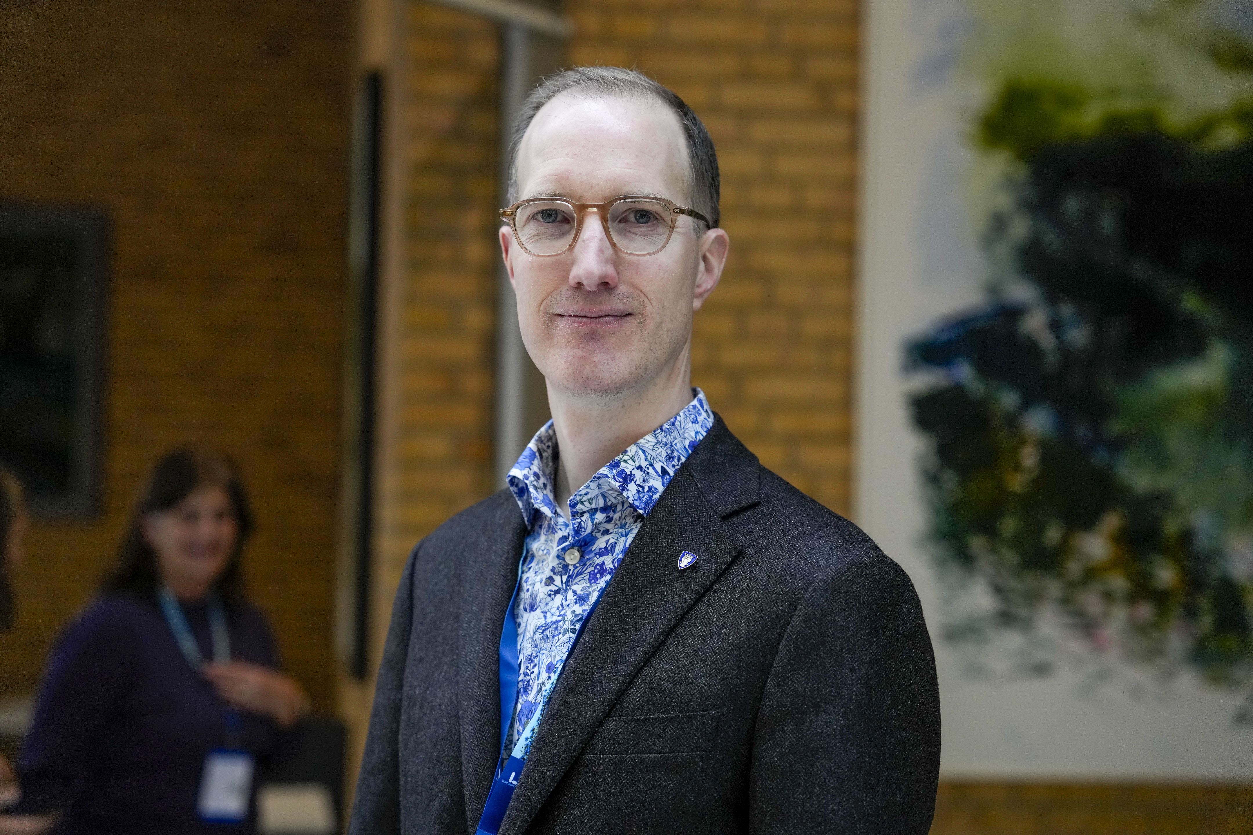 En man med glasögon, mörk kavaj och blåmönstrad skjorta står inomhus med suddig bakgrund av personer och konst.