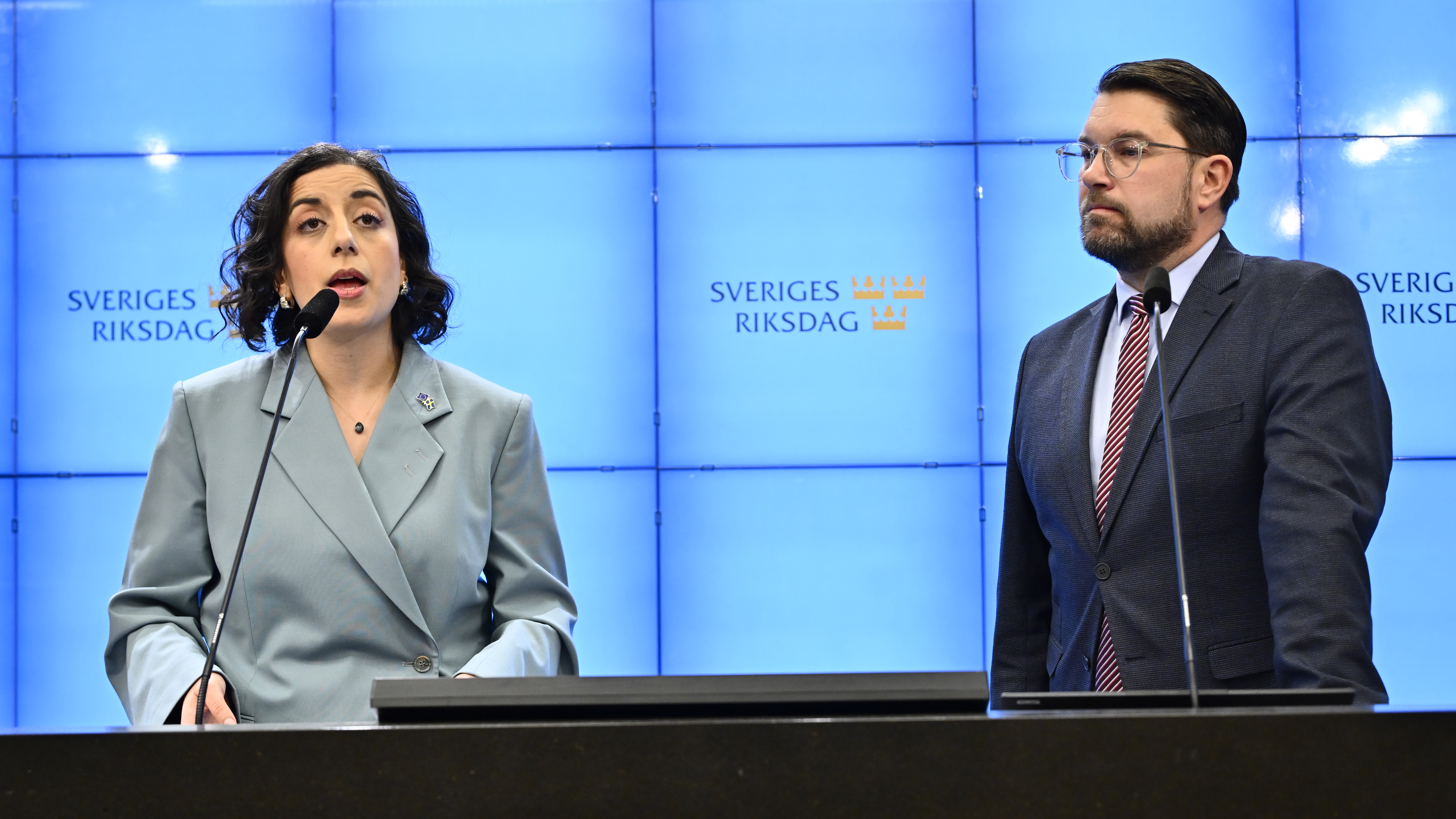 Två personer står vid ett podium framför en blå skärm med Sveriges Riksdags logotyp; en kvinna talar i en mikrofon, medan en man står bredvid.