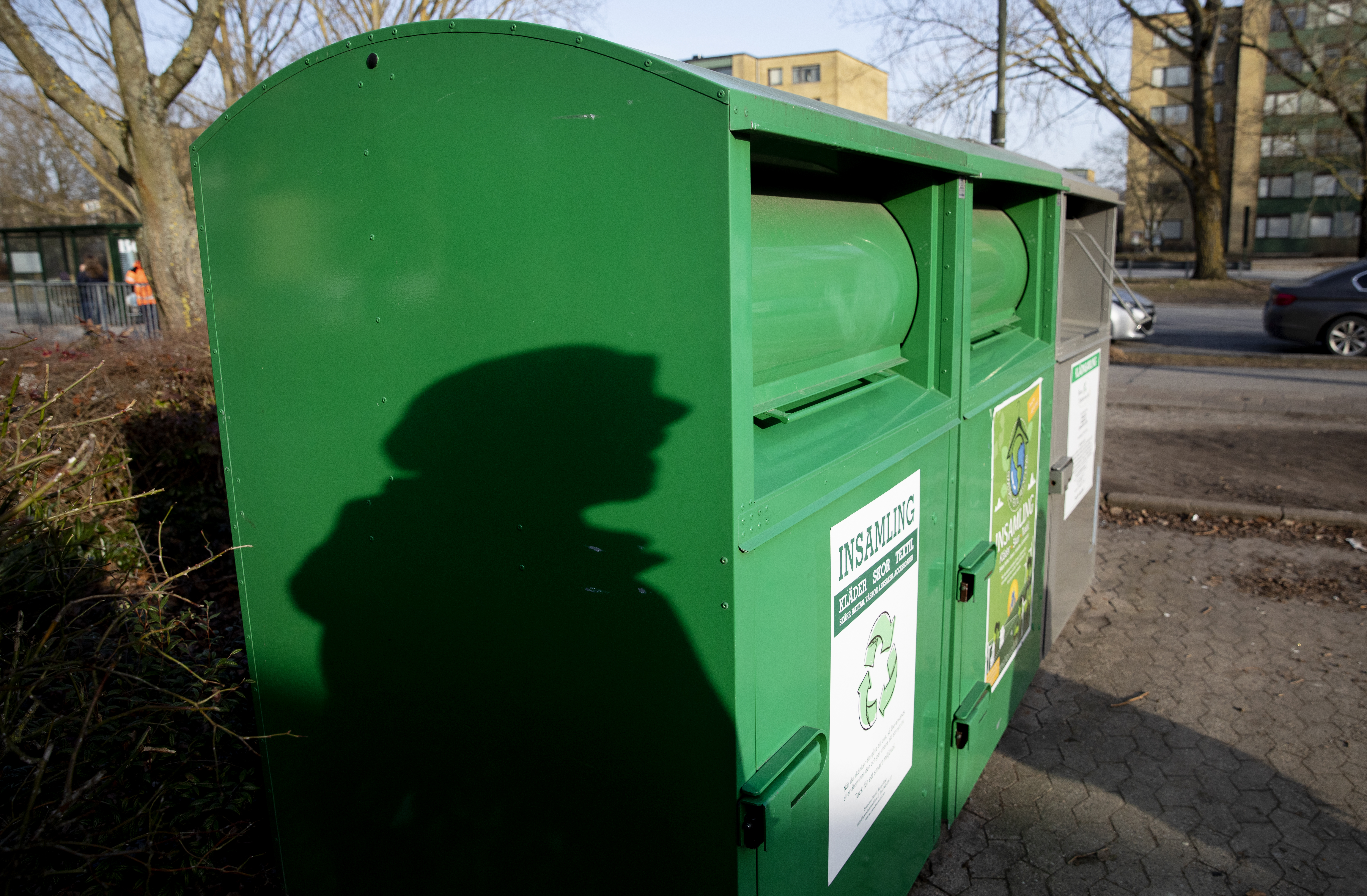 Gröna återvinningskärl utomhus med en skugga av en person på sidan; byggnader och träd syns i bakgrunden.