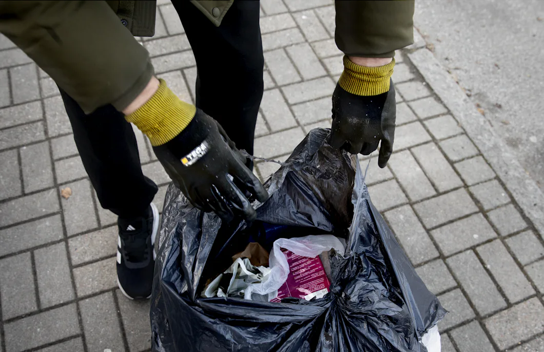 På en asfalterad trottoar slänger en person som bär gula och svarta handskar föremål i svarta soppåsar.