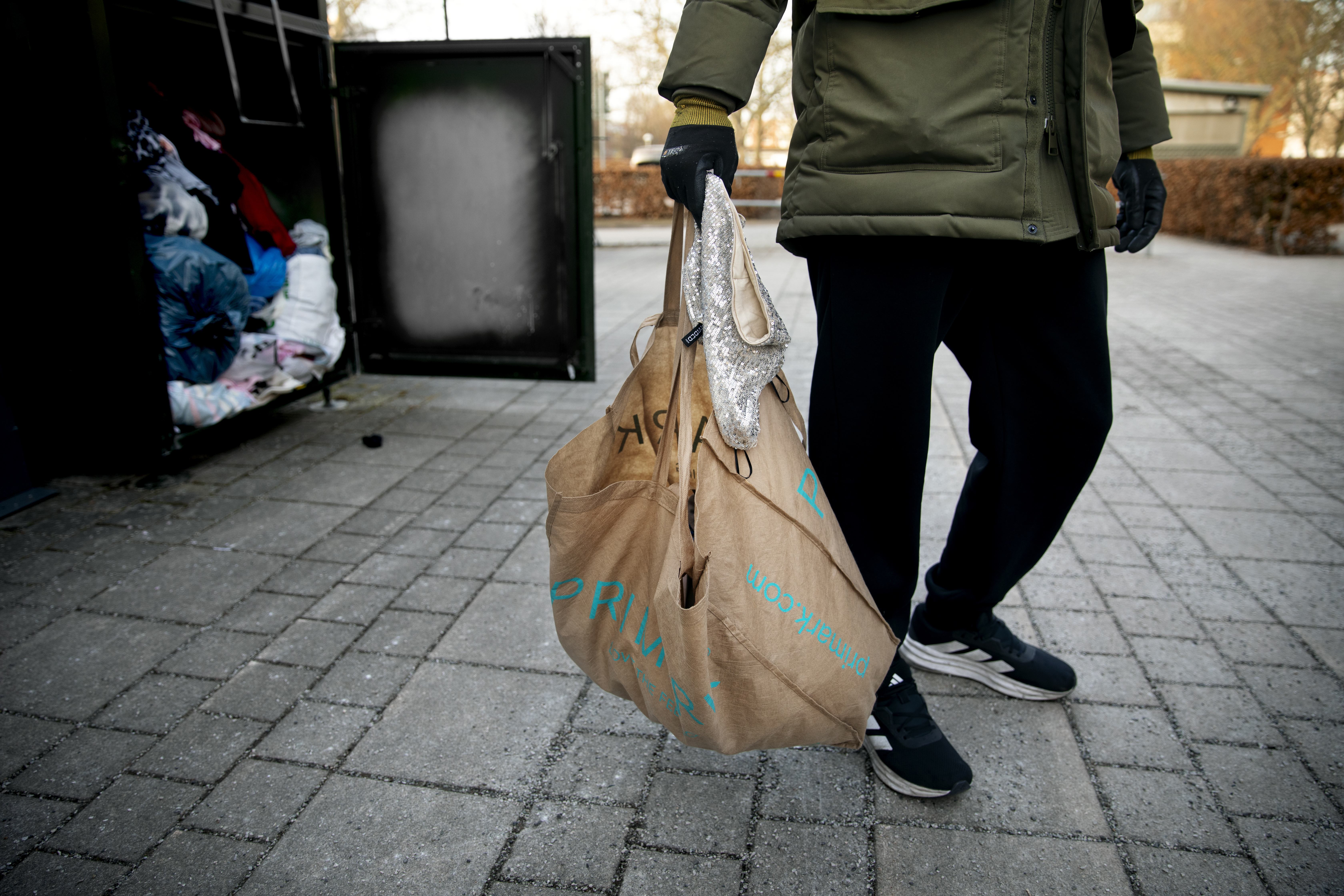 En person med grön jacka och svarta handskar bär en papperspåse nära en öppen utomhusinsamling fylld med kläder.