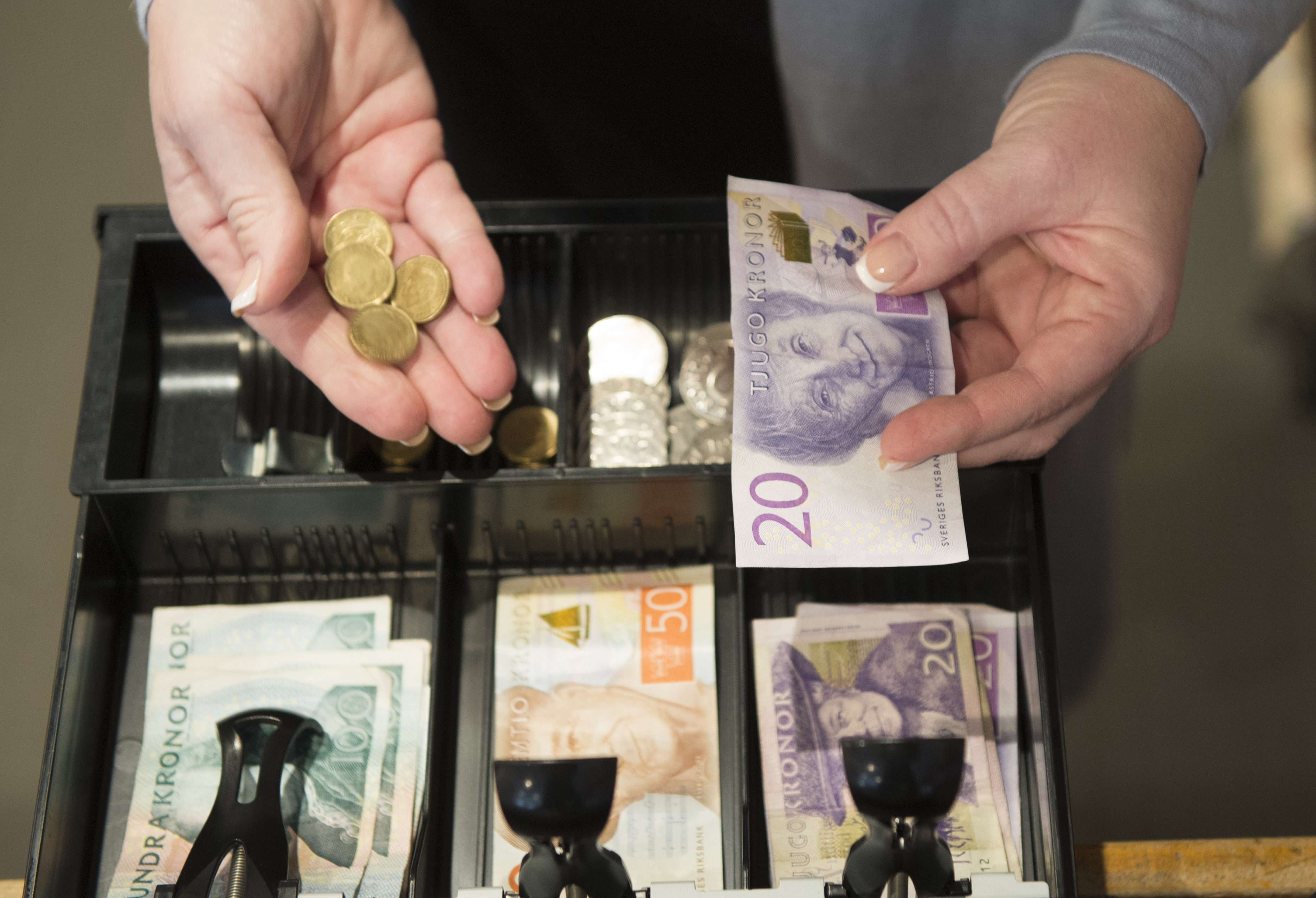 En person håller svenska mynt och en 20-kronorssedel ovanför en öppen kassalåda med svenska sedlar och mynt.