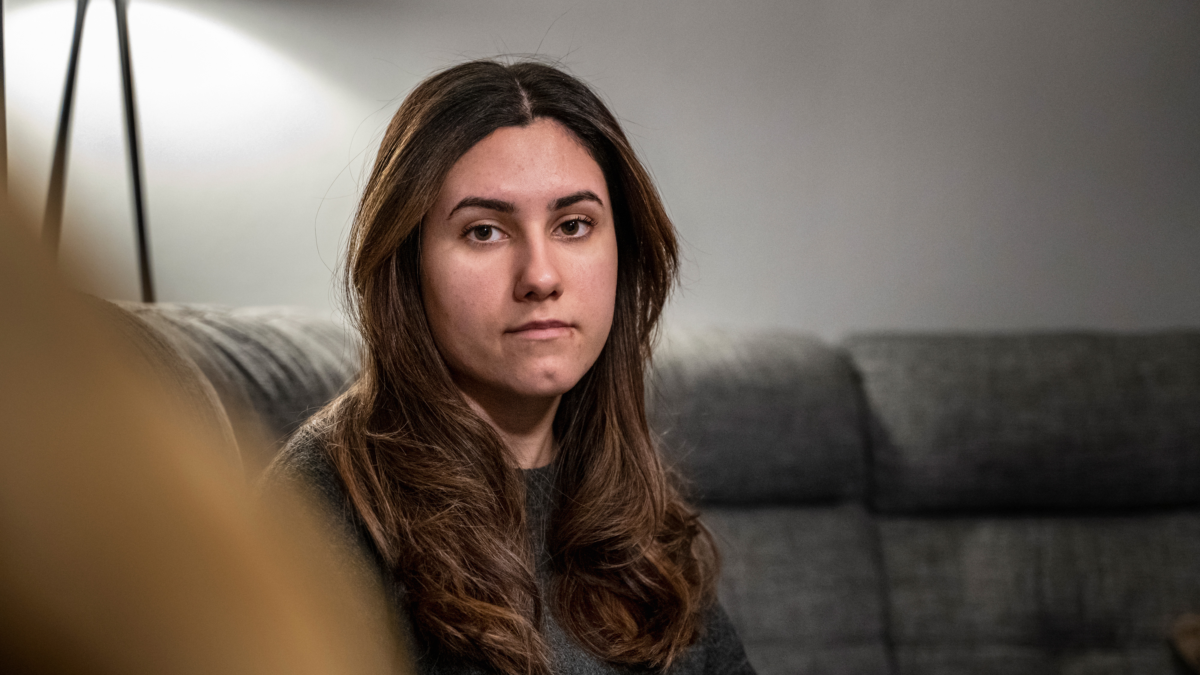 Unga kvinnan Dalya Jaf med långt brunt hår sitter på en grå soffa inomhus och tittar rakt mot kameran med neutral min.