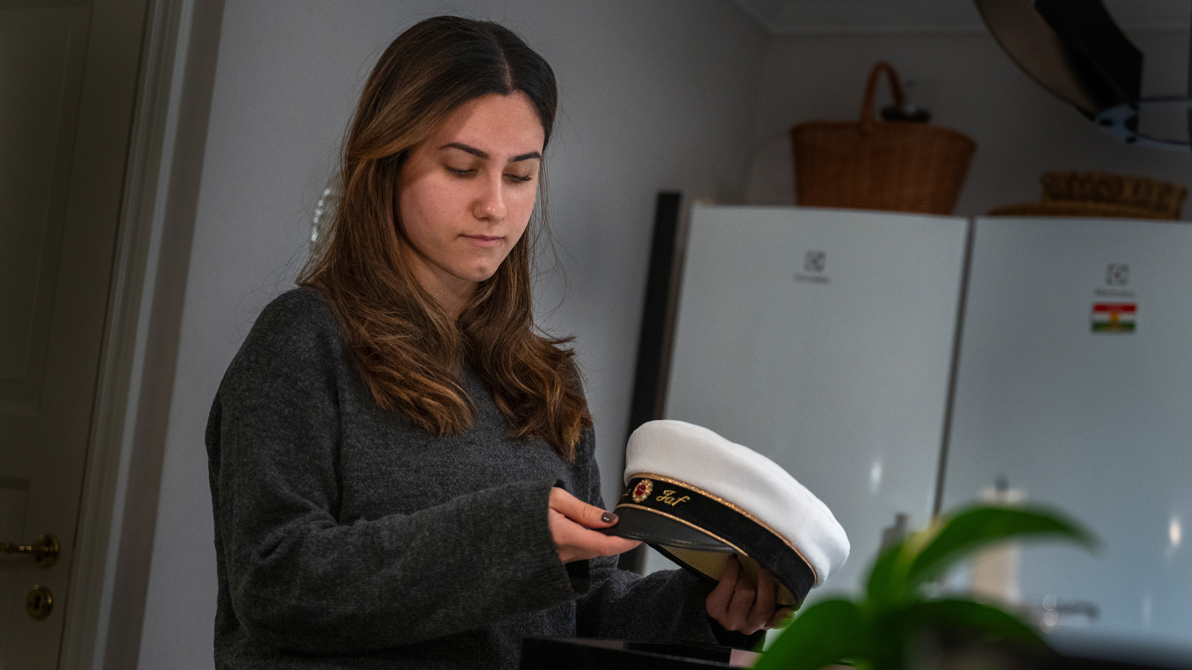 Dalya Jaf med långt brunt hår i en grå tröja håller i en vit studentmössa och står i ett kök.