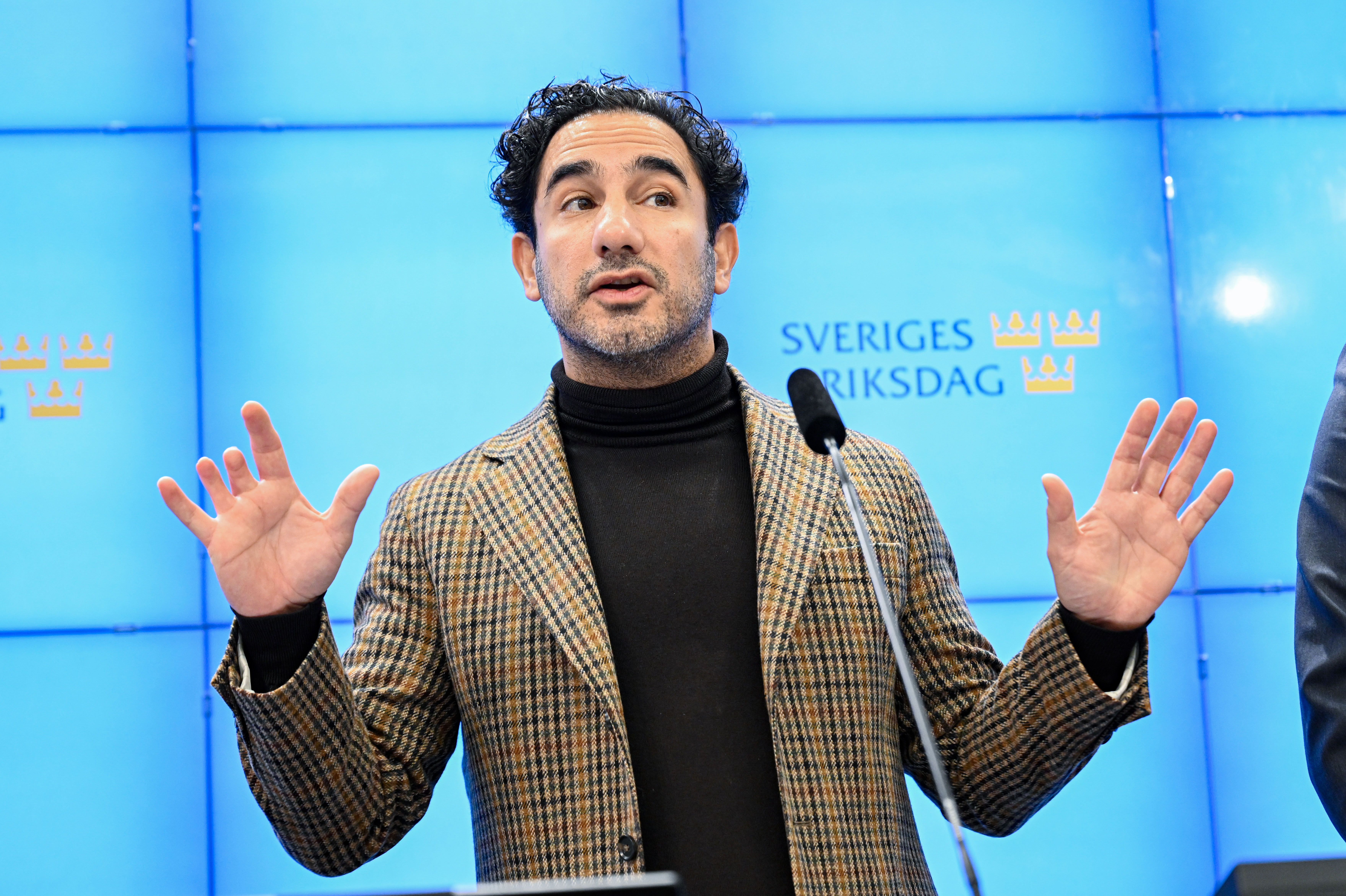 En man i rutig kavaj talar vid ett podium framför en blå bakgrund med Sveriges Riksdags logotyp.