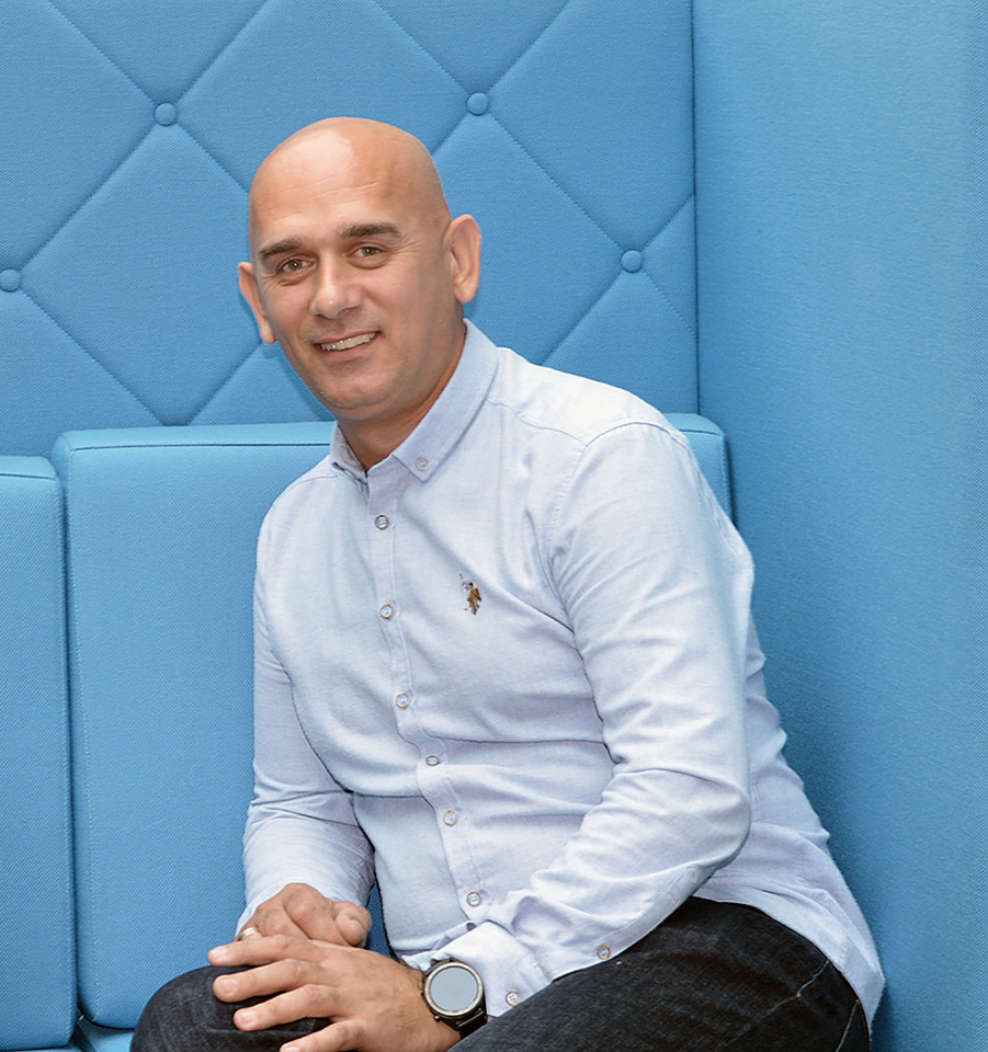 En skallig man med en ljusblå skjorta och mörka byxor sitter på en blå bänk och ler mot kameran.