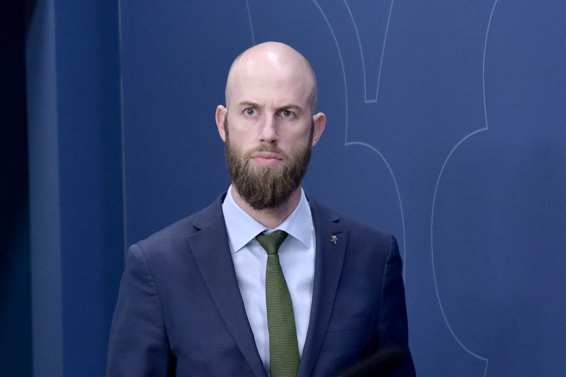 En man med skägg i kostym och gröna slips står framför en blå bakgrund.