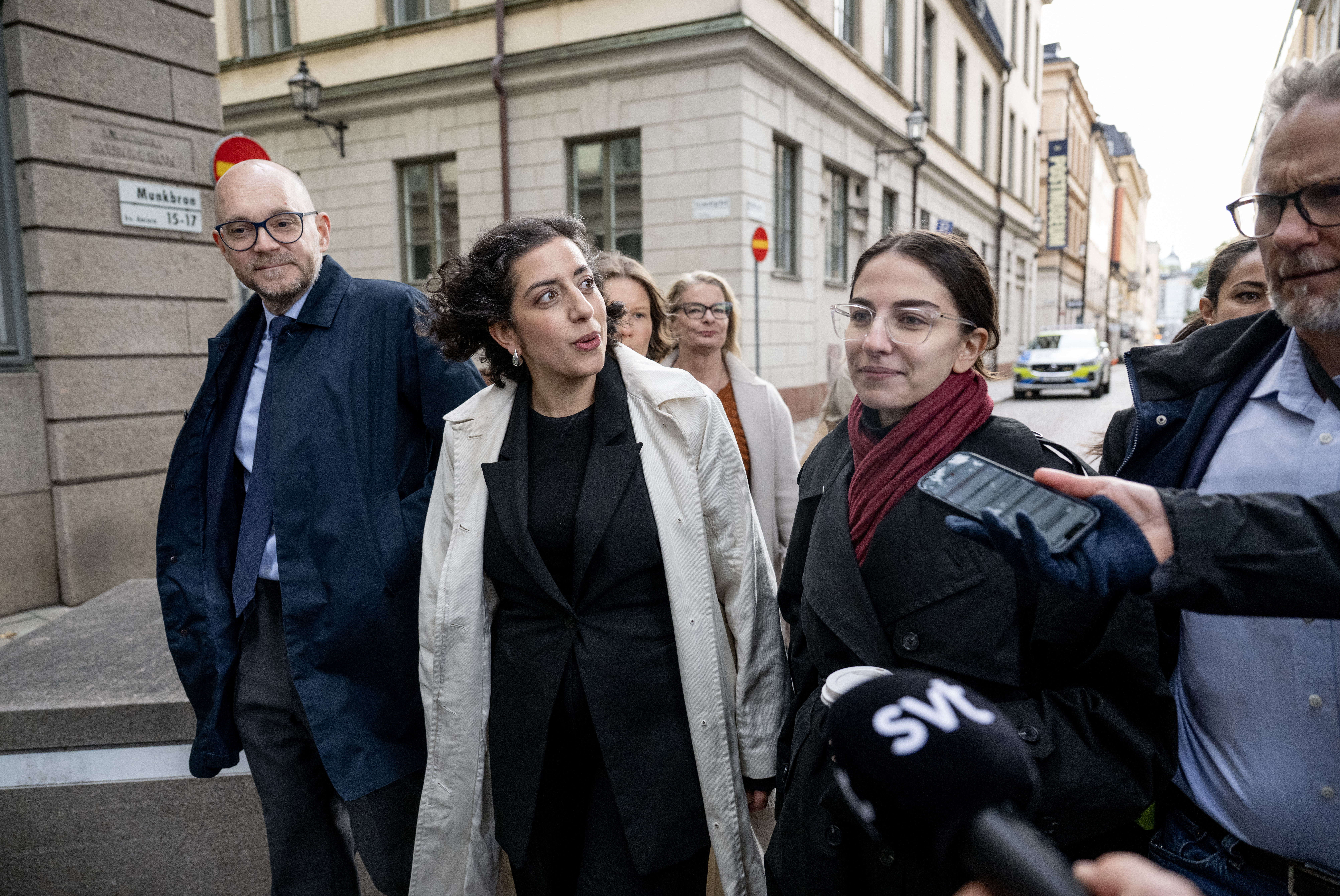 En grupp människor, däribland kvinnor i rockar och sjalar, går på en stadsgata medan reportrar håller fram mikrofoner mot dem.